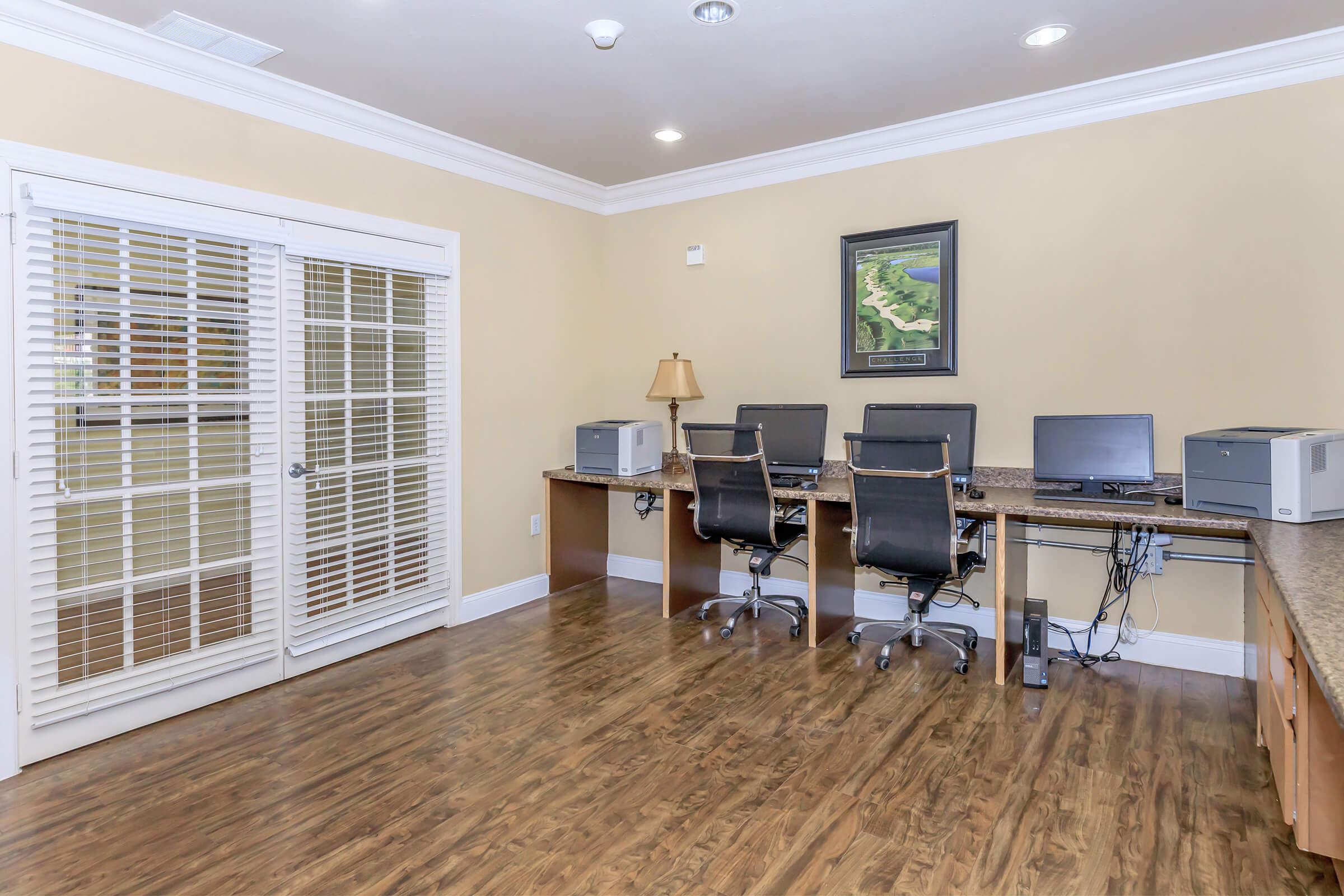 A well-lit office space featuring two desks with computers and printers, wooden flooring, and a framed artwork on the wall. Large windows with blinds allow natural light. A lamp adds additional lighting to the space.