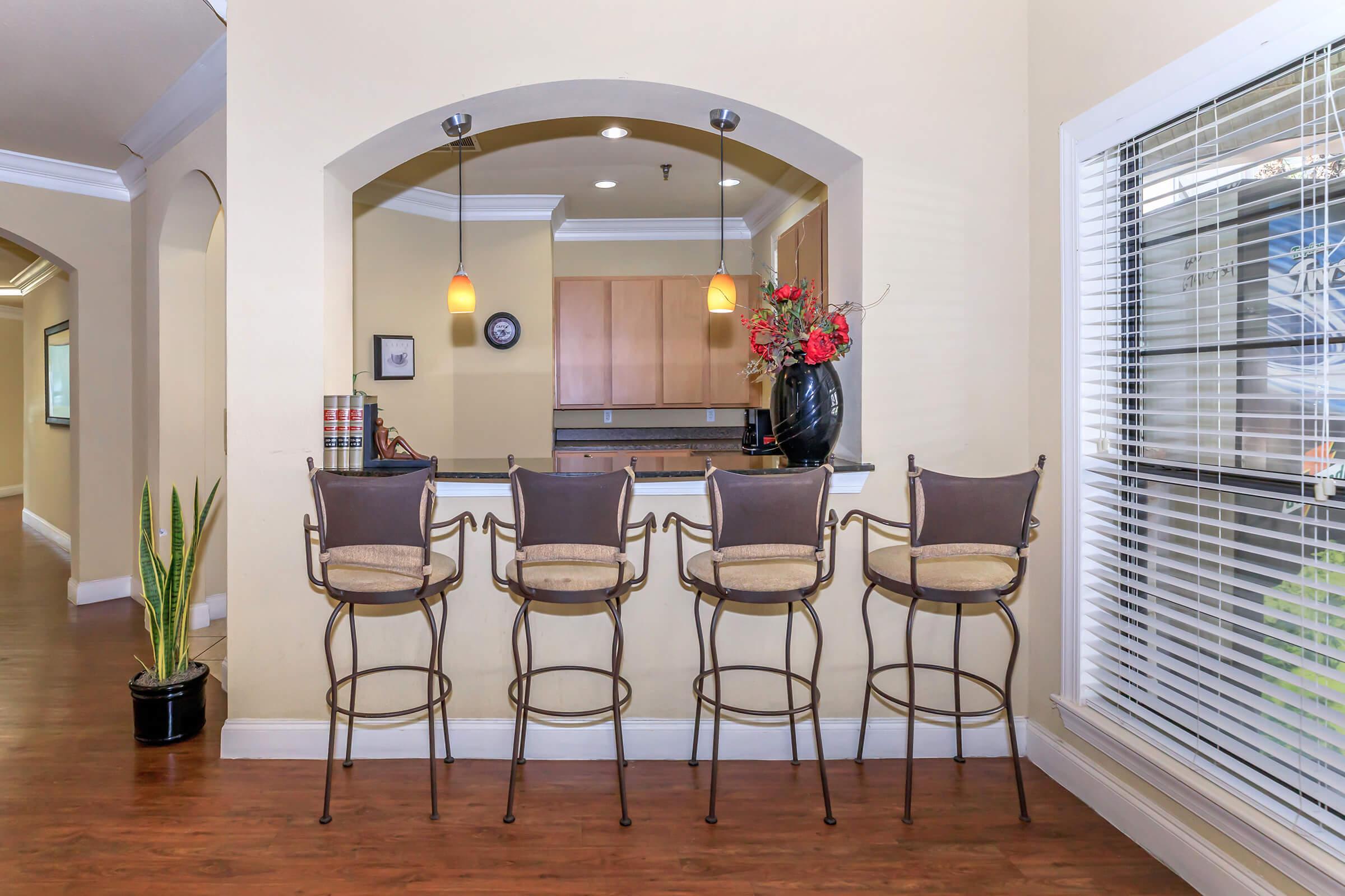A stylish kitchen bar area with four metal stools, a decorative vase with flowers, and pendant lighting. The walls are painted in a warm tone, and there is a window with blinds that provides natural light. The space is inviting and well-decorated, suitable for casual dining or socializing.