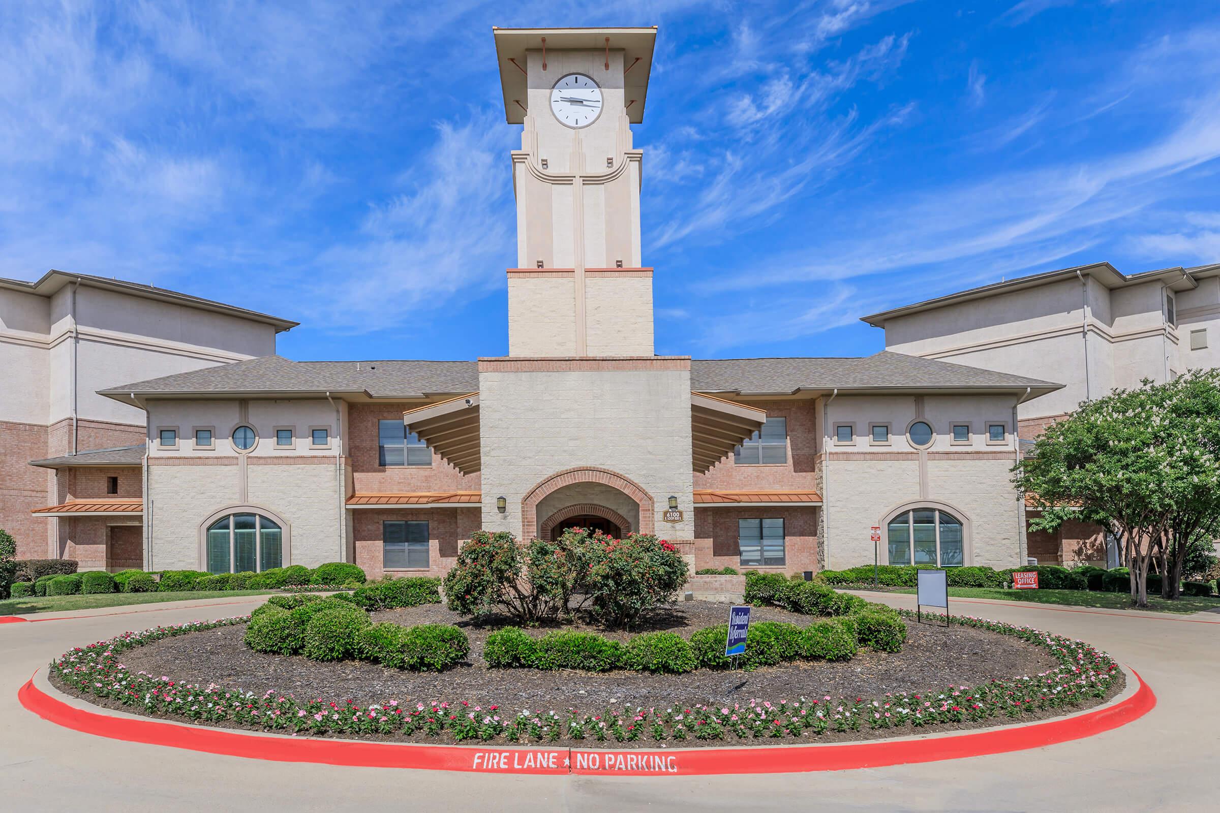 A modern building with a prominent clock tower, surrounded by manicured landscaping. The entrance features a circular drive with a red "FIRE LANE - NO PARKING" sign. The sky is blue with a few clouds, creating a bright and welcoming atmosphere.