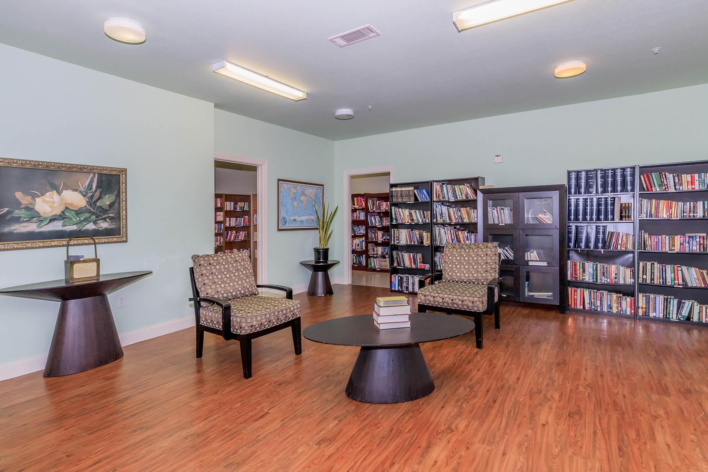 A cozy reading room featuring two patterned armchairs and a circular coffee table. The walls are painted light green, and there are bookshelves filled with books in the background. A framed painting of flowers is displayed on one wall, and a small plant is placed on a table. The room is well-lit and inviting.