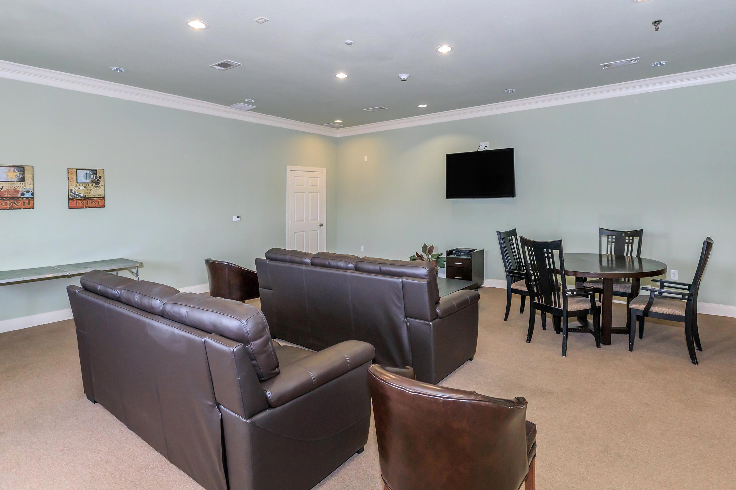 Interior view of a spacious lounge area featuring two dark leather sofas, a wooden dining table with six chairs, and a flat-screen TV mounted on a light green wall. The carpeted floor adds warmth to the room, which is well-lit and has minimal decorative elements.