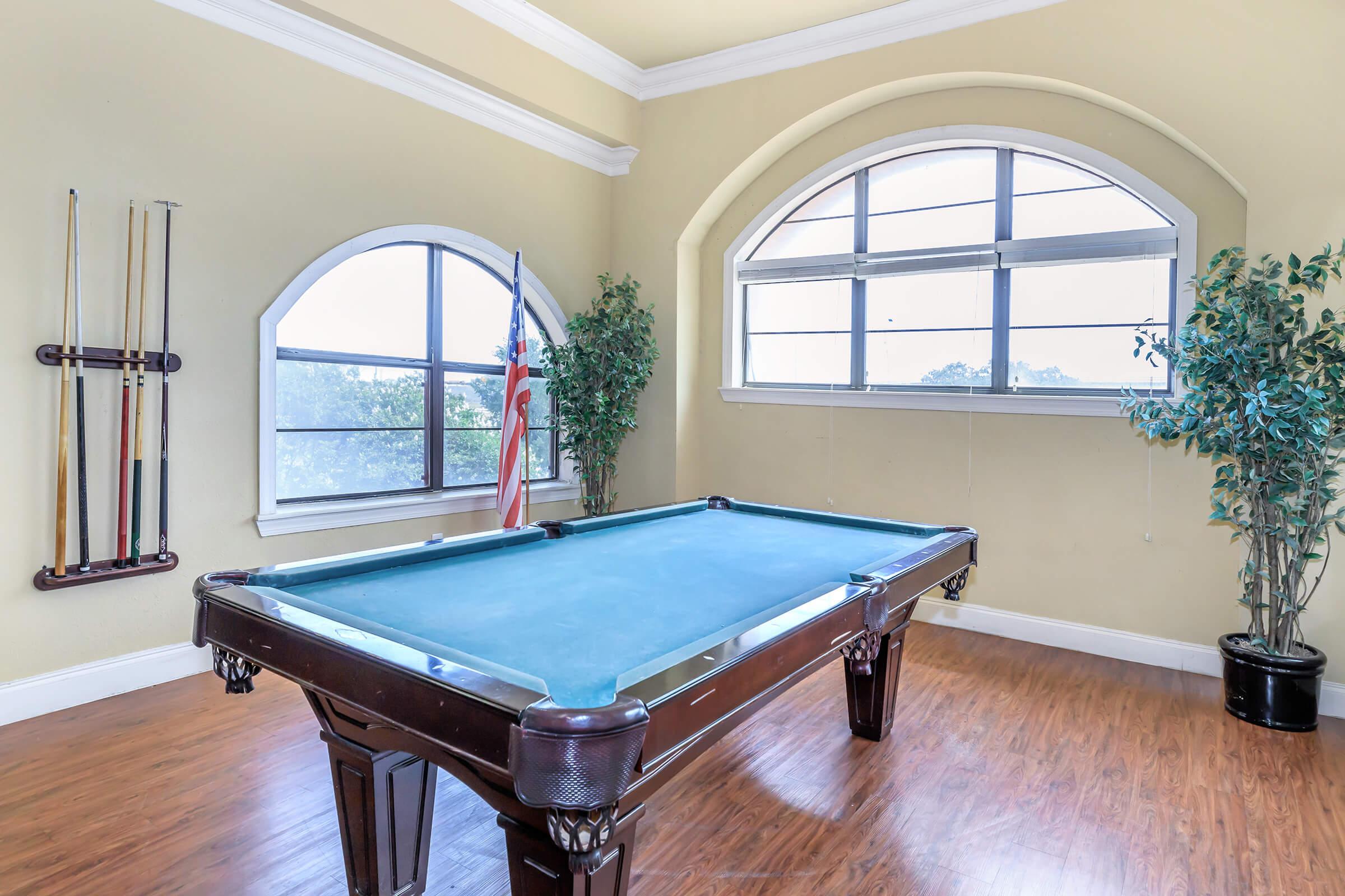 A well-lit room featuring a green felt pool table with a polished wooden frame. Large windows with arching tops provide natural light, and an American flag is positioned by the window. A plant in a pot adds a touch of greenery, while cue sticks are mounted on the wall. The flooring is a warm wood laminate.