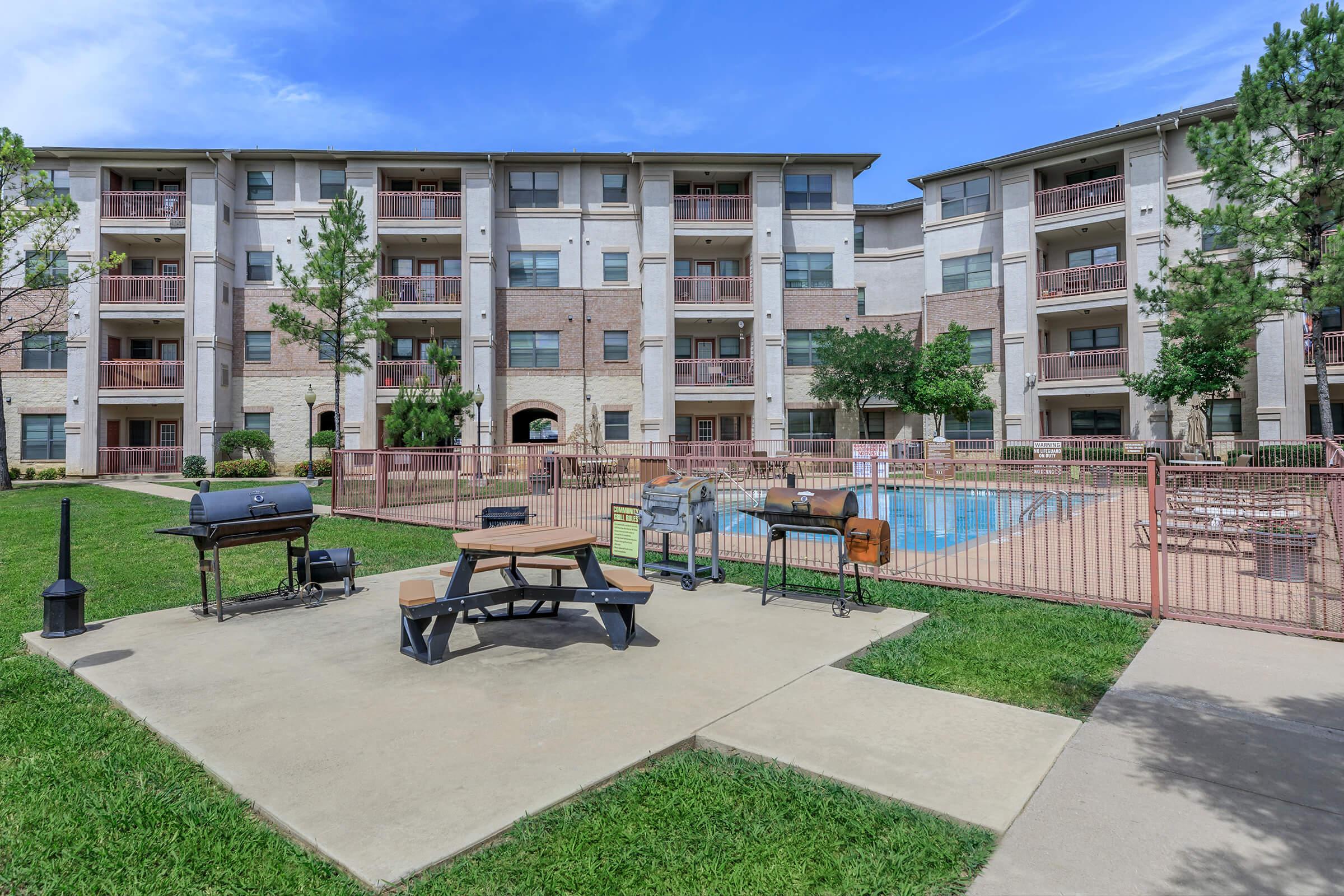 A multi-story apartment building with balconies and a swimming pool in front. There is a picnic table and a grill set up on a concrete patio. Lush green grass surrounds the area, and trees add to the outdoor ambiance. The sky is blue with a few clouds.