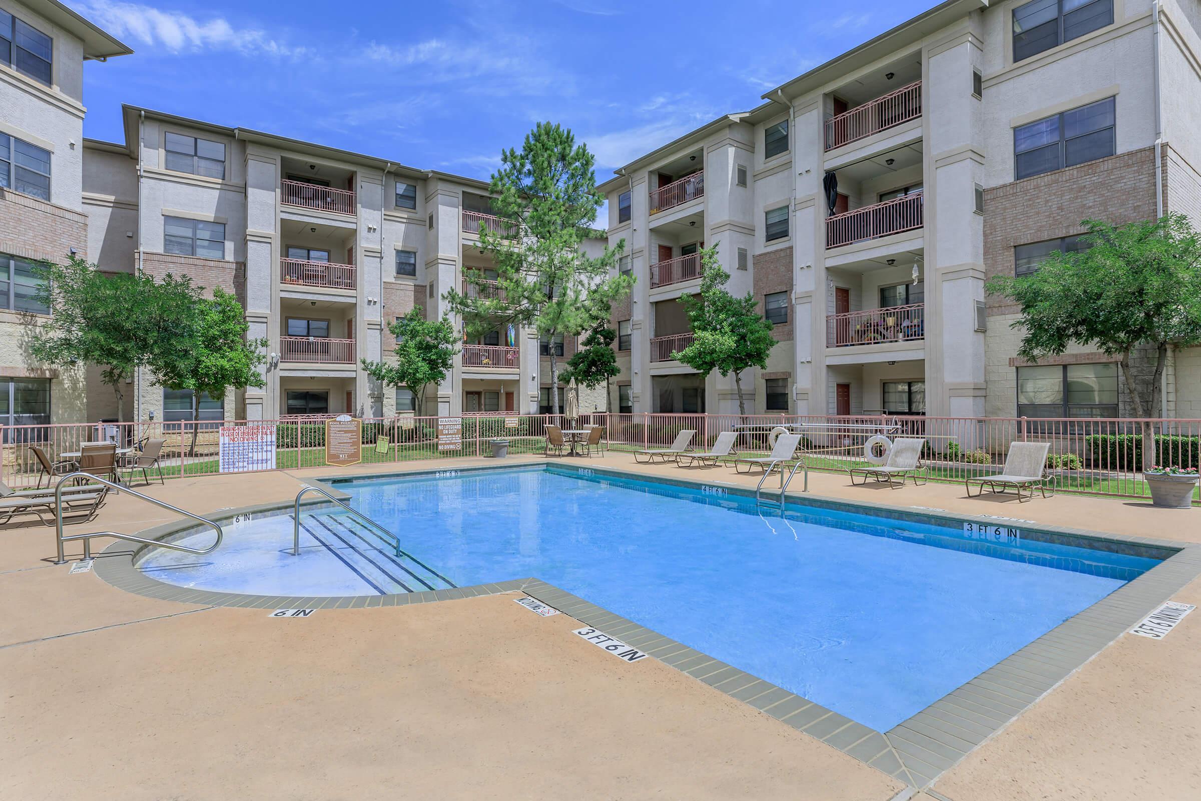 A serene apartment complex courtyard featuring a sparkling blue swimming pool surrounded by lounge chairs. The pool area is framed by four-story residential buildings, green trees, and well-maintained landscaping under a clear blue sky.