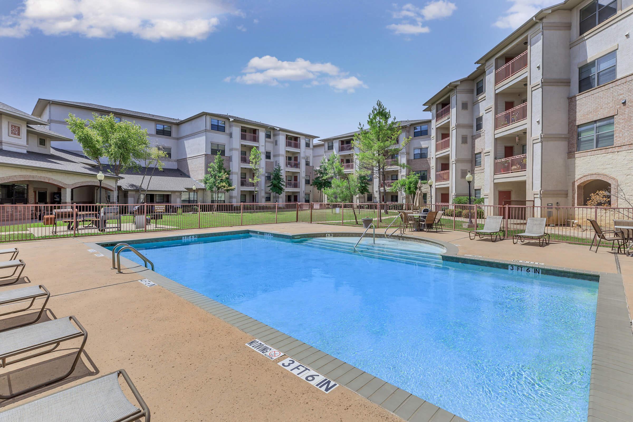 A clear blue swimming pool surrounded by lounge chairs, with lush green landscaping and apartment buildings in the background. Bright blue sky with a few clouds completes the scene, creating a relaxing poolside atmosphere.