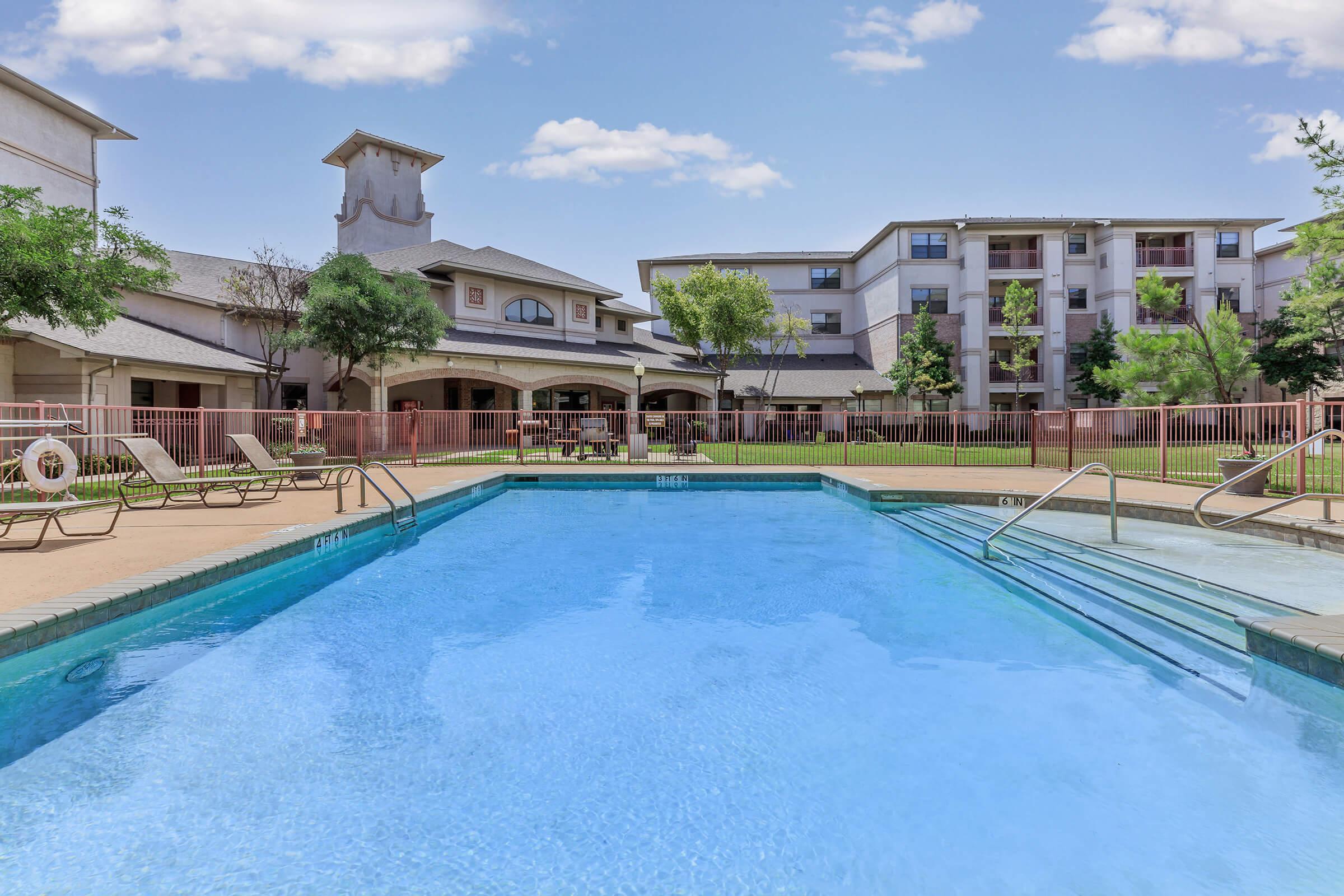 A clear blue swimming pool surrounded by lounge chairs, with a well-maintained grassy area in the background. There are two buildings in the distance, and trees providing shade. The sky is sunny with a few clouds, creating a relaxing outdoor atmosphere.