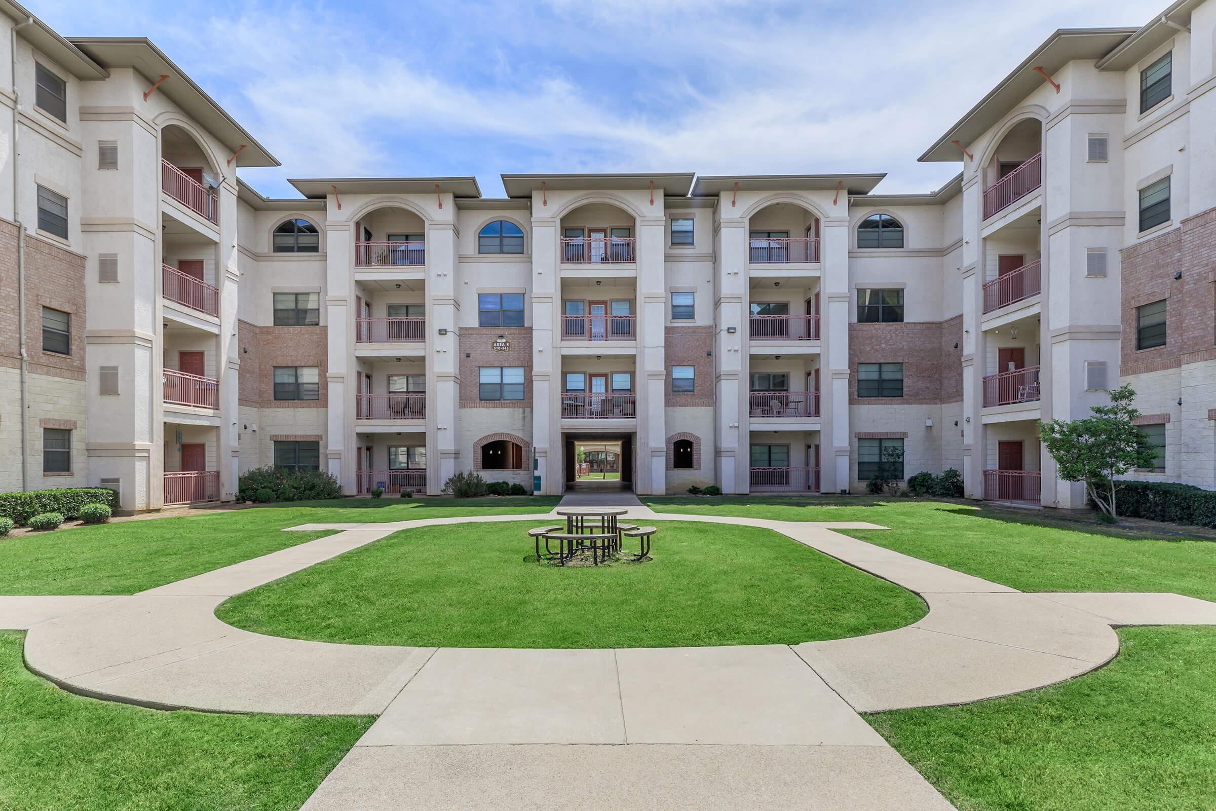 An image of a modern multi-story apartment building, featuring a central green area with landscaped lawns and sidewalks. The building has balconies on each floor, with large windows and a light-colored facade. Blue sky with some clouds can be seen above.
