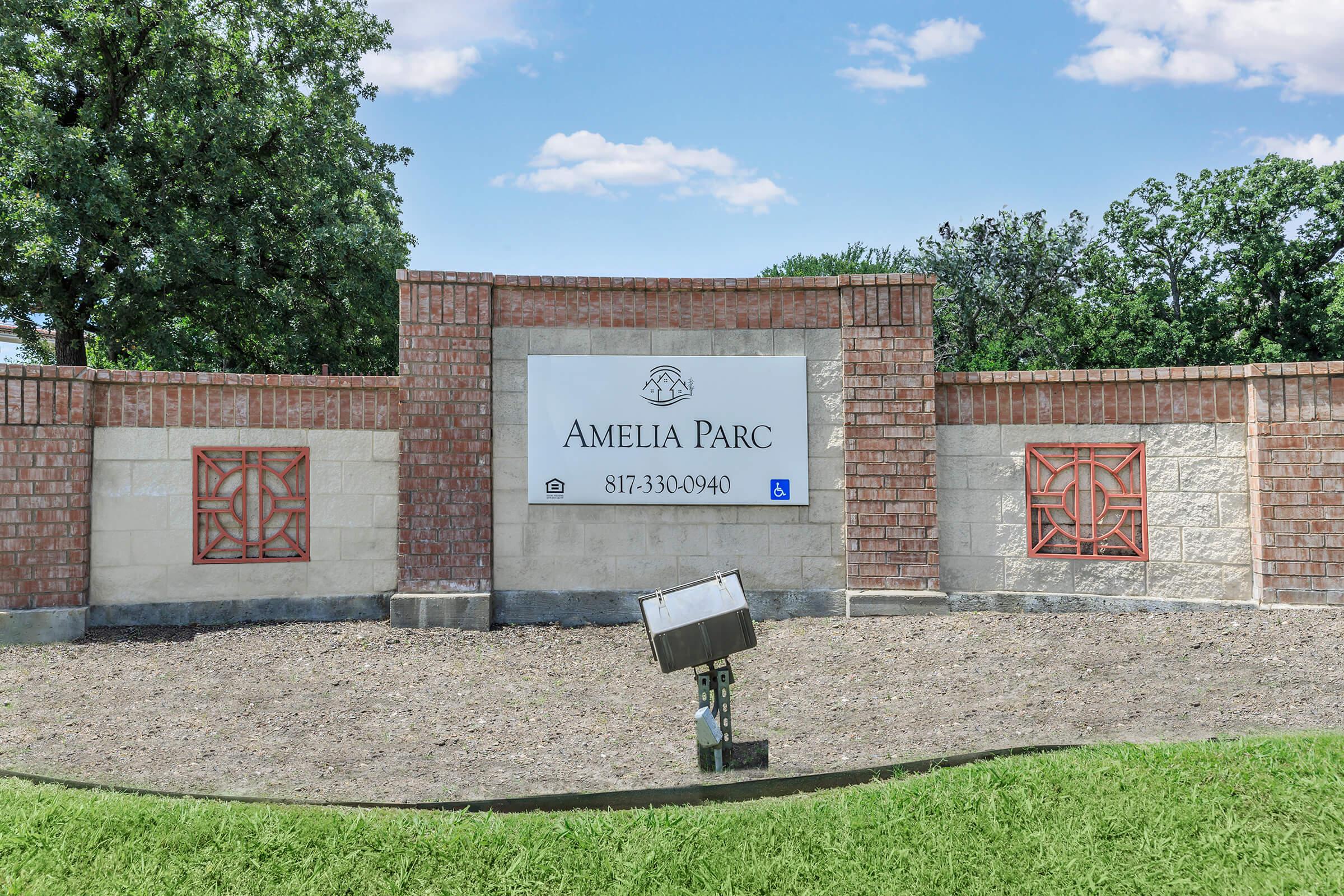 Sign for Amelia Parc with contact number 817-330-0940, featuring a brick and stone structure with decorative elements. The background includes greenery and a clear blue sky with a few clouds.