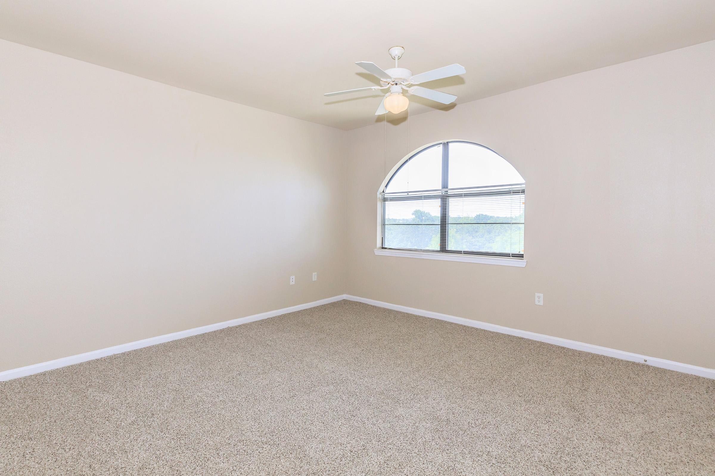 Spacious, empty room with light beige walls and carpet flooring. A large arched window allows natural light to enter, featuring white window blinds. The ceiling is equipped with a modern ceiling fan, enhancing the room’s airy feel. Ideal for various furniture arrangements and decorating options.