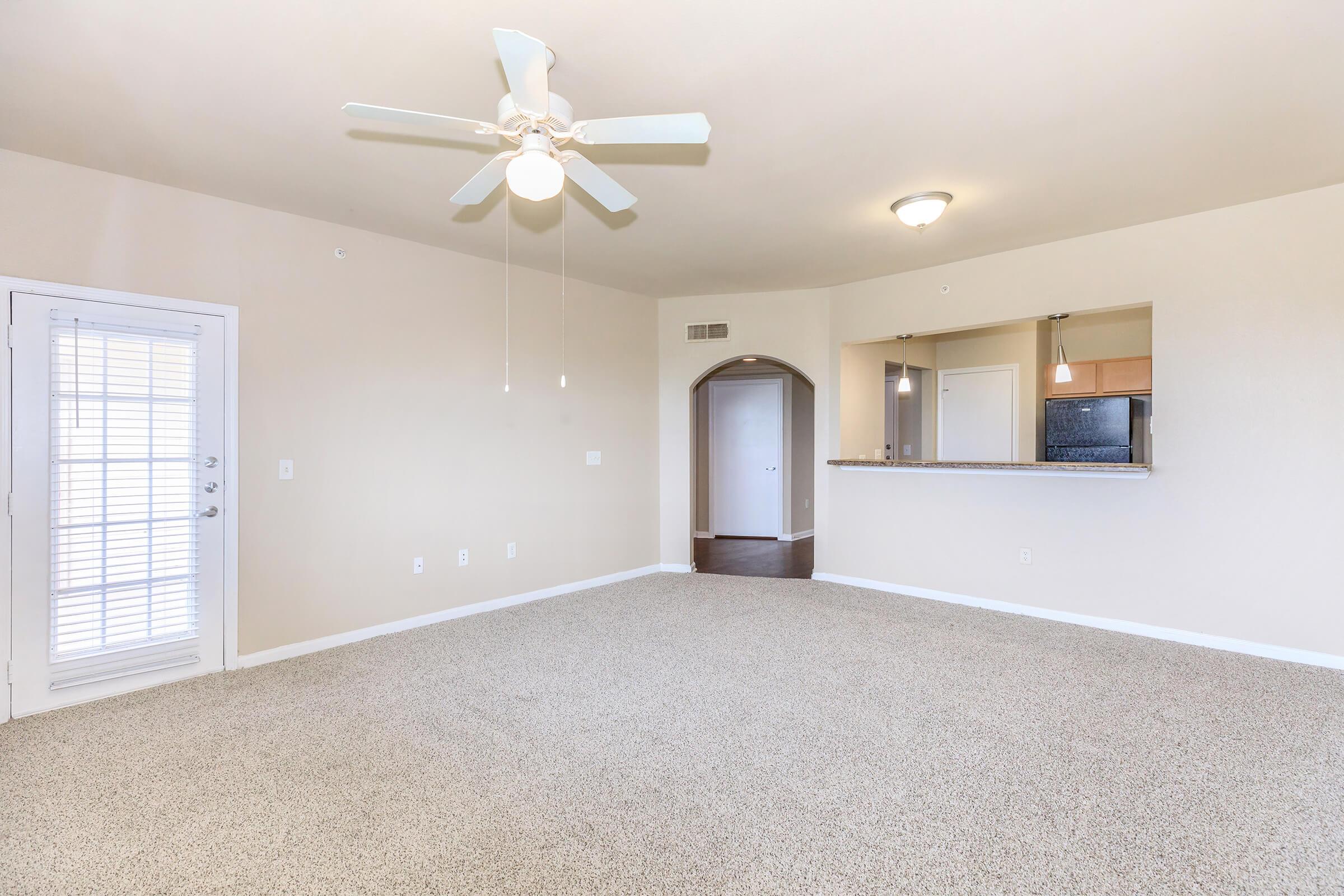 A spacious, light-colored living area featuring a ceiling fan, beige walls, and carpeted flooring. There is a doorway leading to a kitchen area with a breakfast bar. Natural light streams in through a window with blinds, and a door opens to a small patio or balcony.