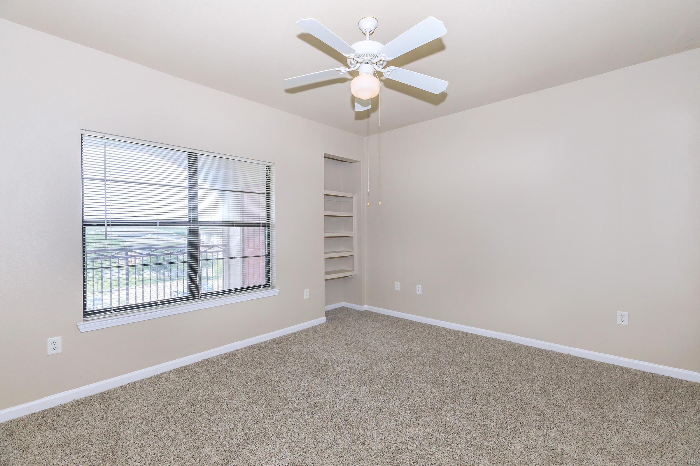 A spacious, empty room with light-colored walls and carpeted flooring. It features a ceiling fan, a large window with blinds allowing natural light, and built-in shelves on one wall. The overall atmosphere is bright and neutral, making it suitable for various purposes.