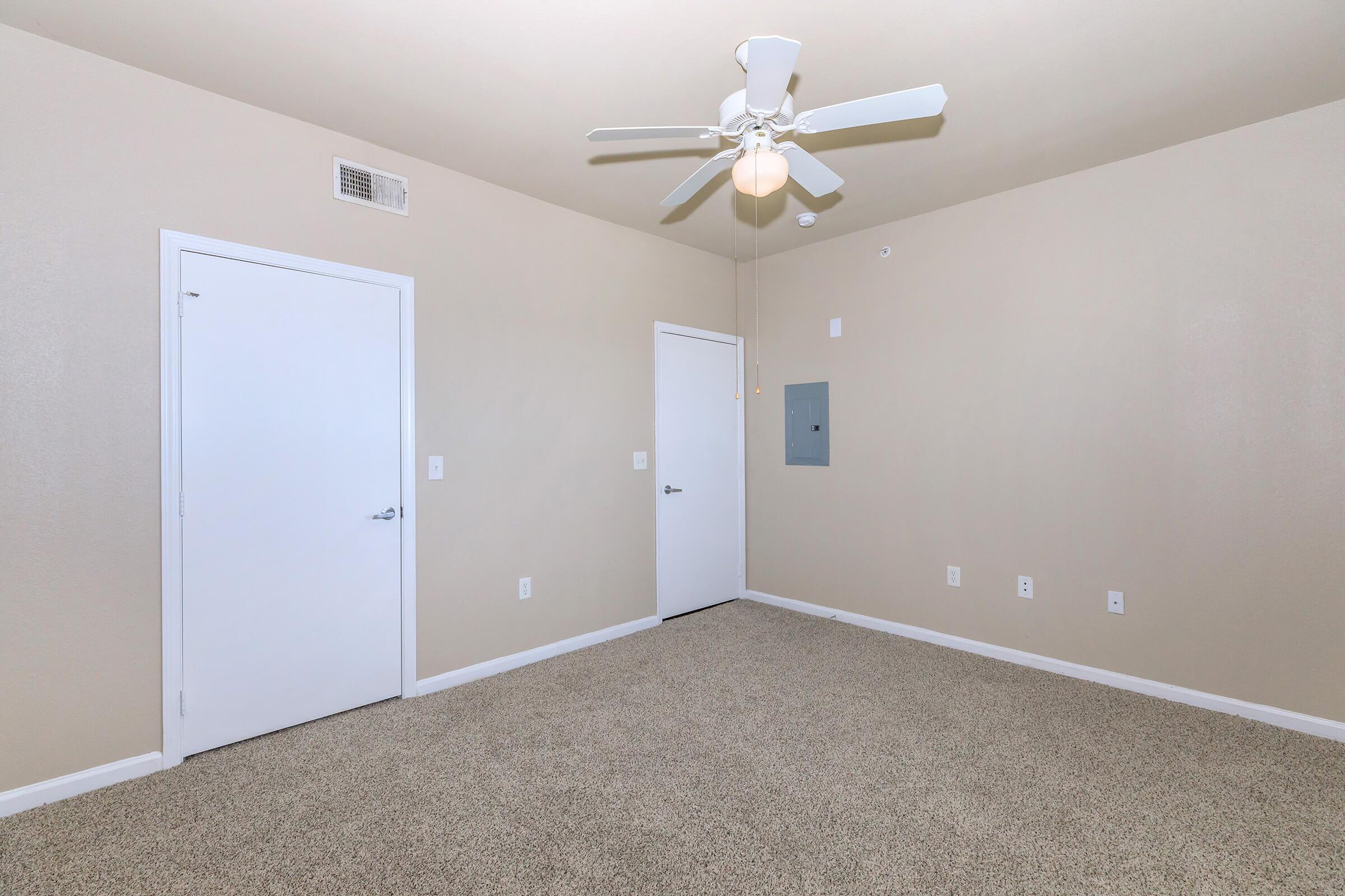 Empty room featuring light beige walls, a ceiling fan with a light fixture, and two white doors. The floor is covered in beige carpet, and there are minimal wall fixtures. The room has a clean, spacious feel with plenty of natural light.