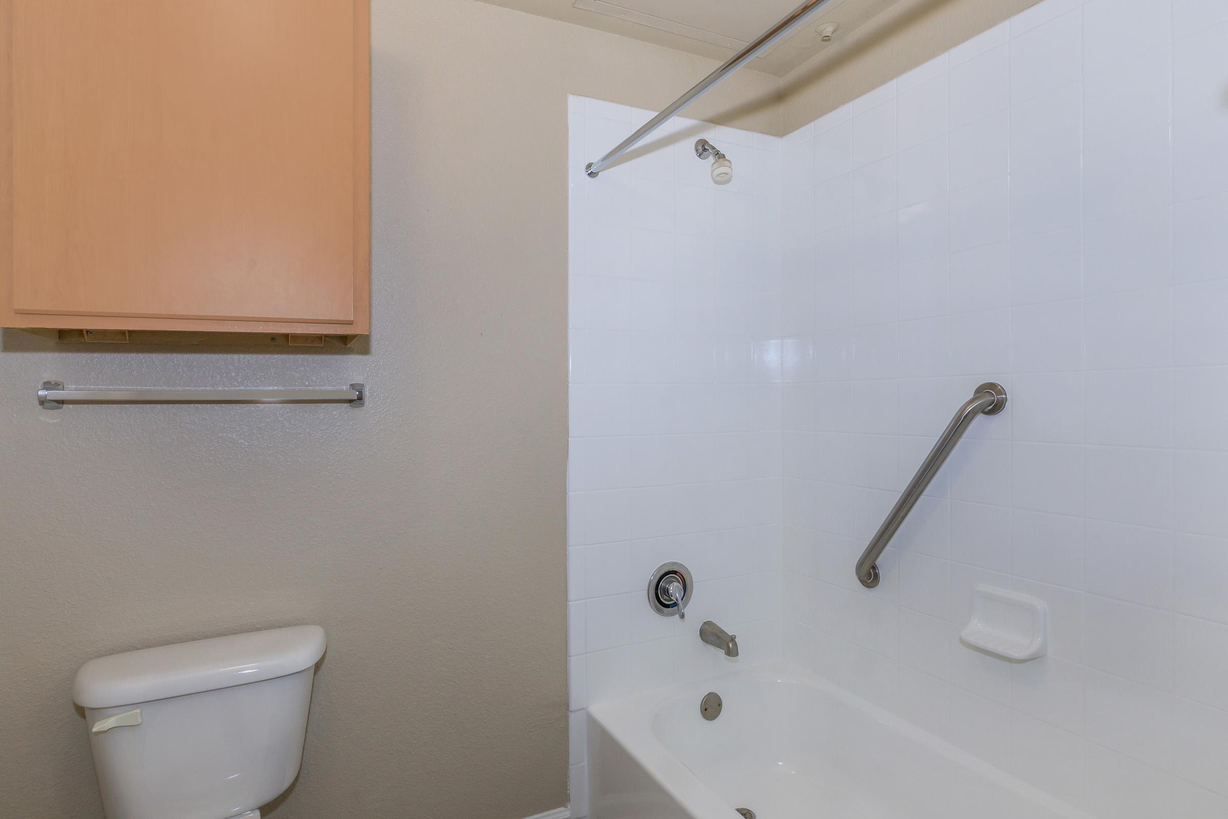 A clean bathroom featuring a white bathtub with a shower, a wall-mounted grab bar, and a toilet nearby. The walls are painted a light color, and there is a wooden cabinet above the bathtub for storage. The overall appearance is modern and functional.