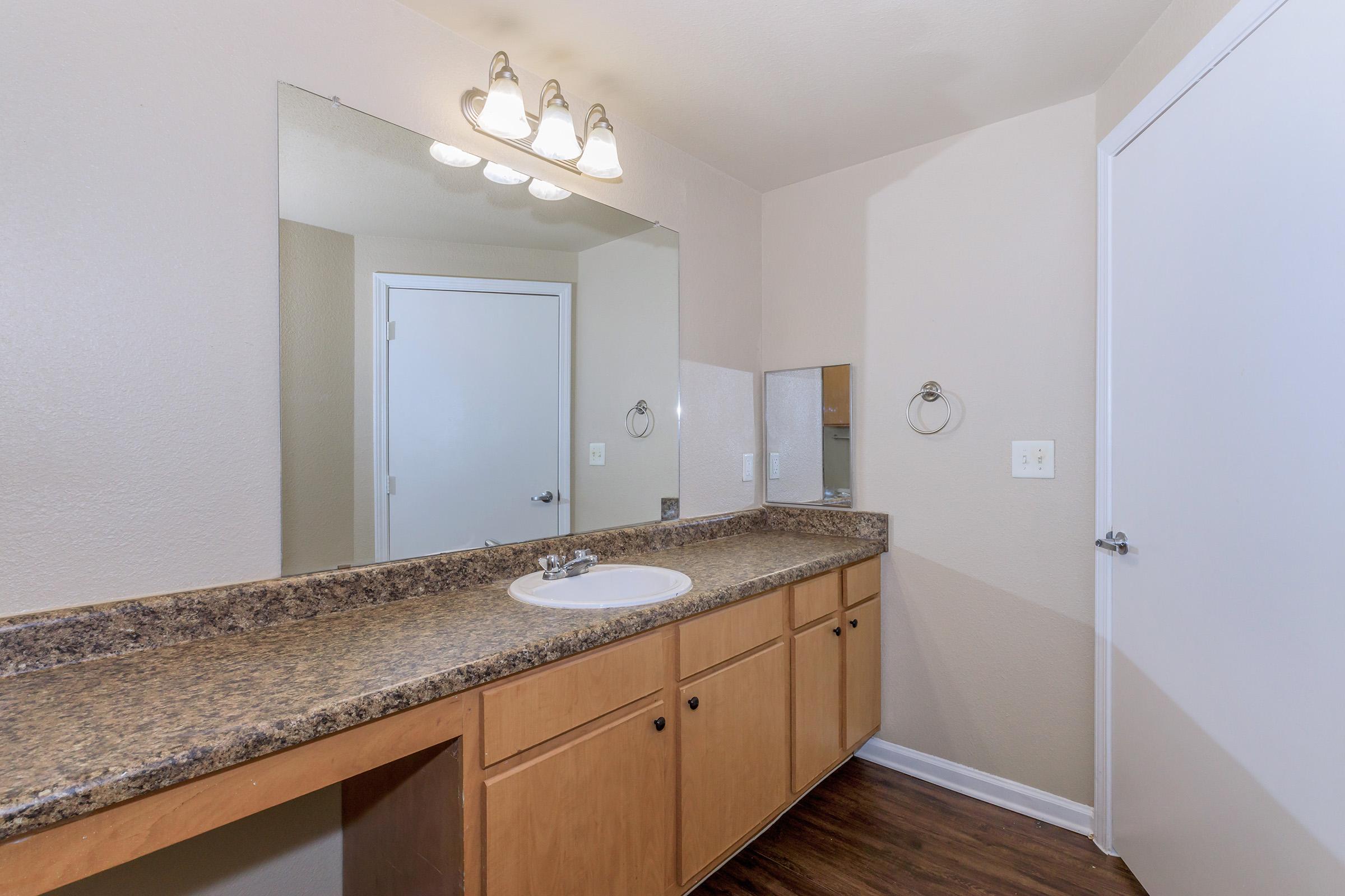 A clean, modern bathroom with a large mirror above a granite countertop. The wood cabinets provide storage, and there is a white sink. A light fixture with three bulbs is mounted above the mirror. A door leads to another room, and the walls are painted in a neutral color, enhancing the spacious feel.