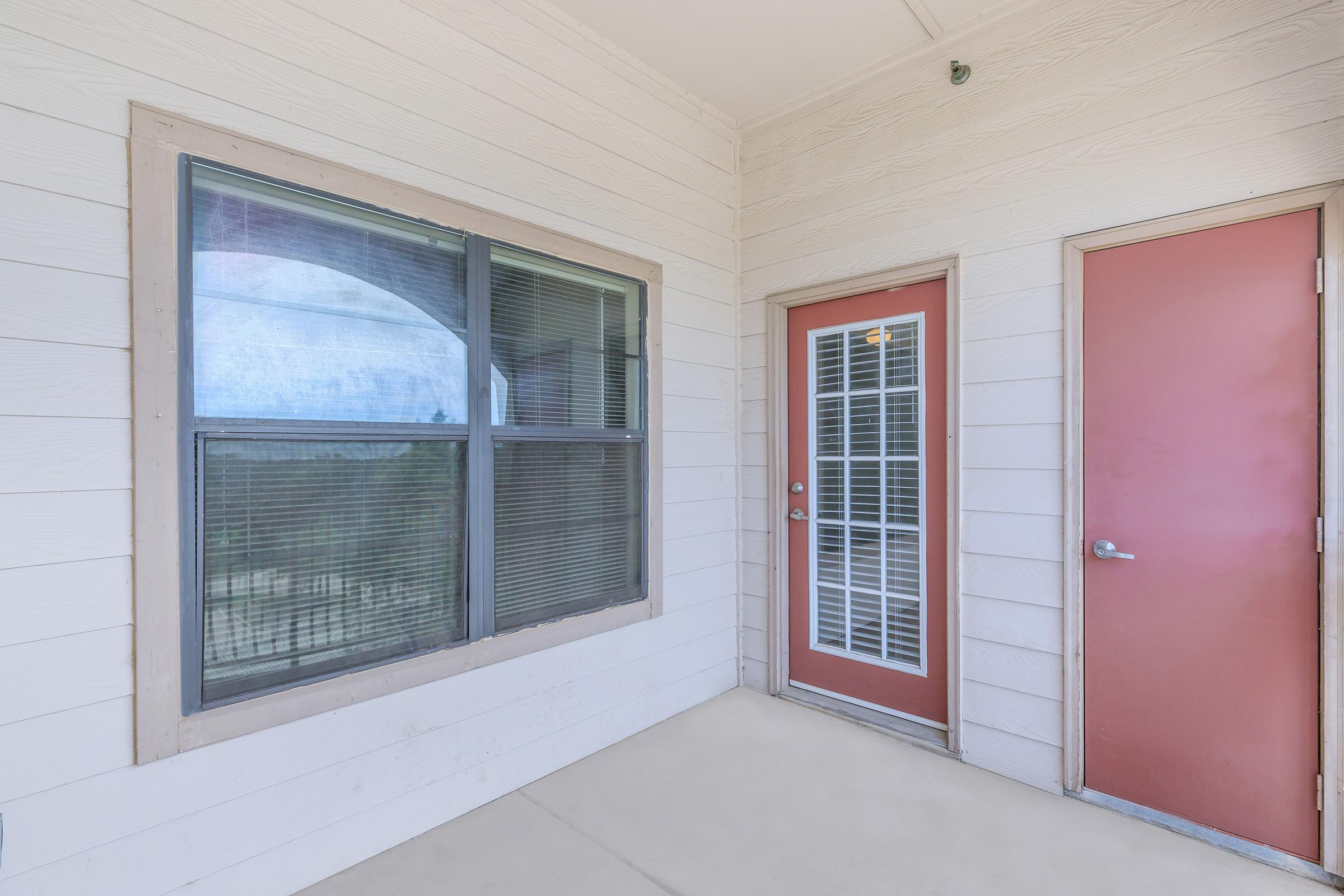 A view of a small exterior porch area featuring a door with a glass panel and a window with grid patterns. The walls are light-colored with a textured finish, and the floor is concrete. The scene shows a simple and modern entrance to a building.