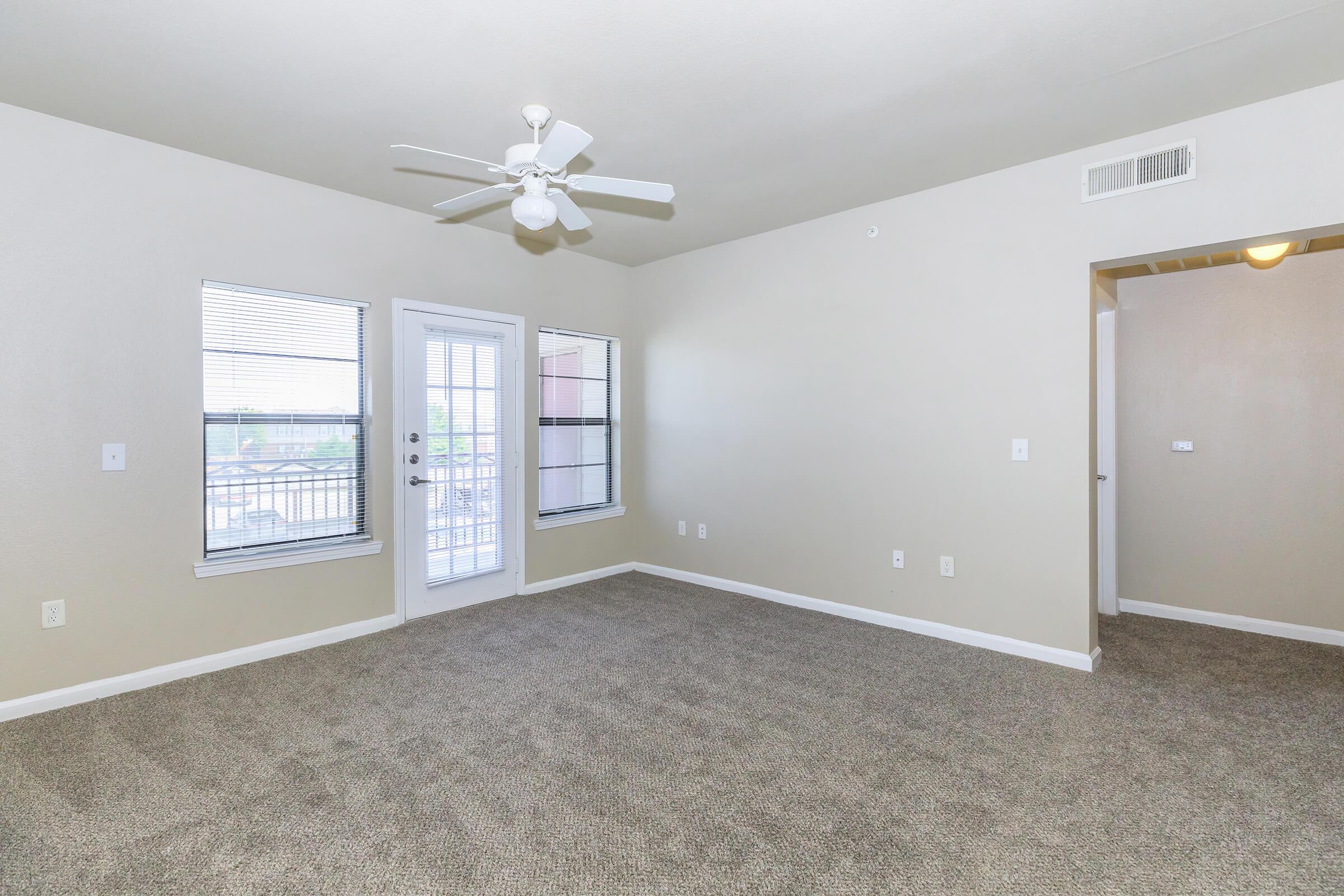 Empty living room with light beige walls and carpeted flooring. Features large windows allowing natural light, a ceiling fan, and a glass door leading to a balcony. Room has minimal décor and electrical outlets on the walls.
