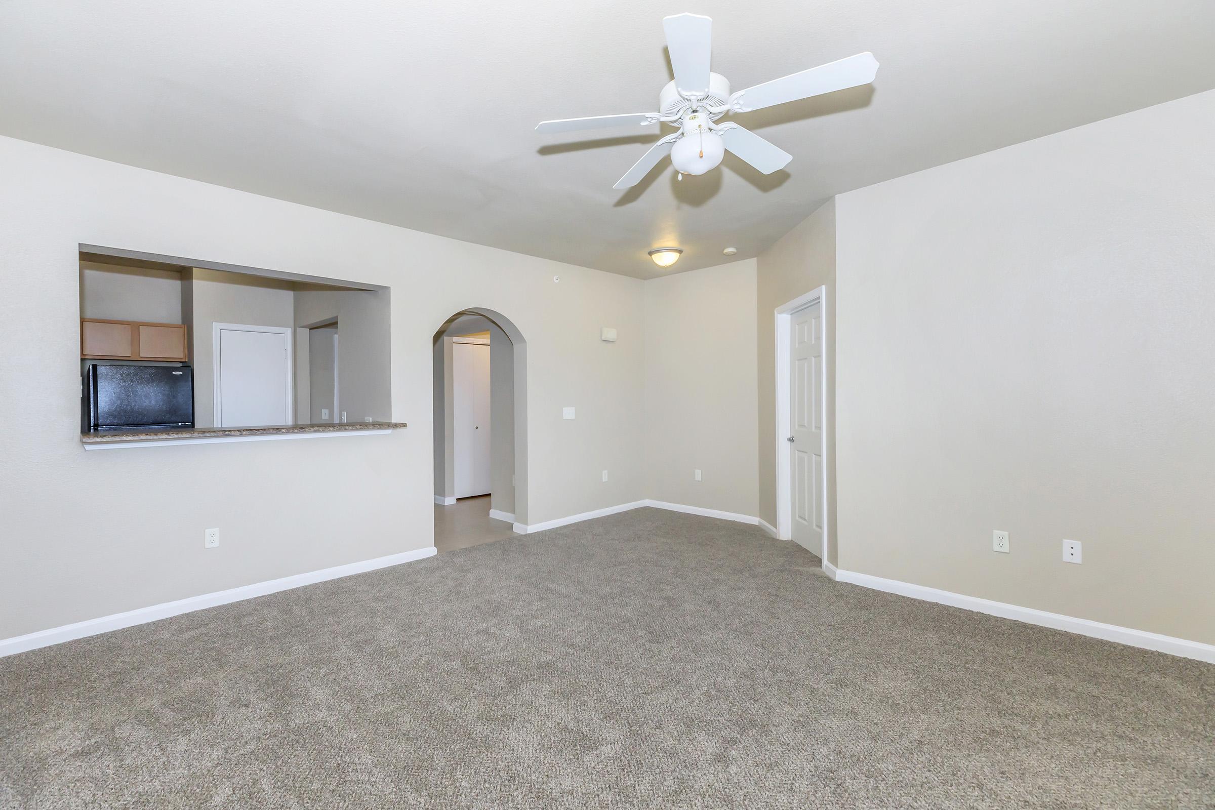 A spacious, light-colored living room featuring a ceiling fan, beige carpet, and neutral walls. An open archway leads to another room, while a bar area connects to a kitchen with wooden cabinets. The room is well-lit, with a door visible on the right and a large window for natural light.