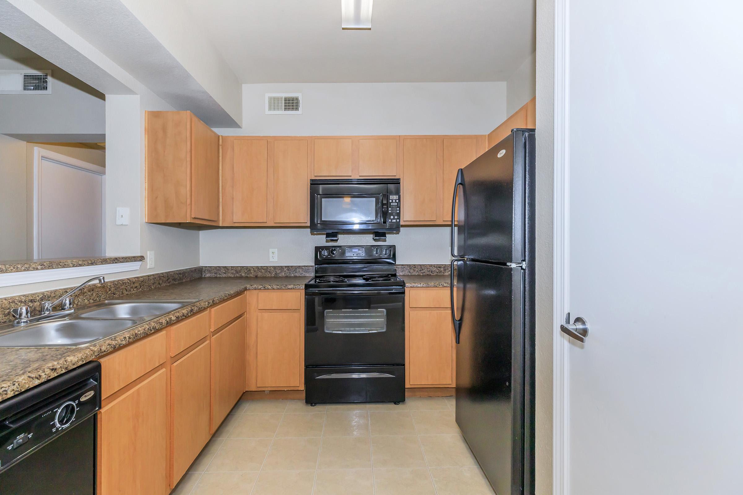 Modern kitchen featuring wood cabinets, granite countertops, a black stove and microwave, and a black refrigerator. The space has a neutral tile floor and ample lighting from a ceiling fixture. There are two sinks with an adjacent counter area, providing a functional layout for cooking and meal preparation.