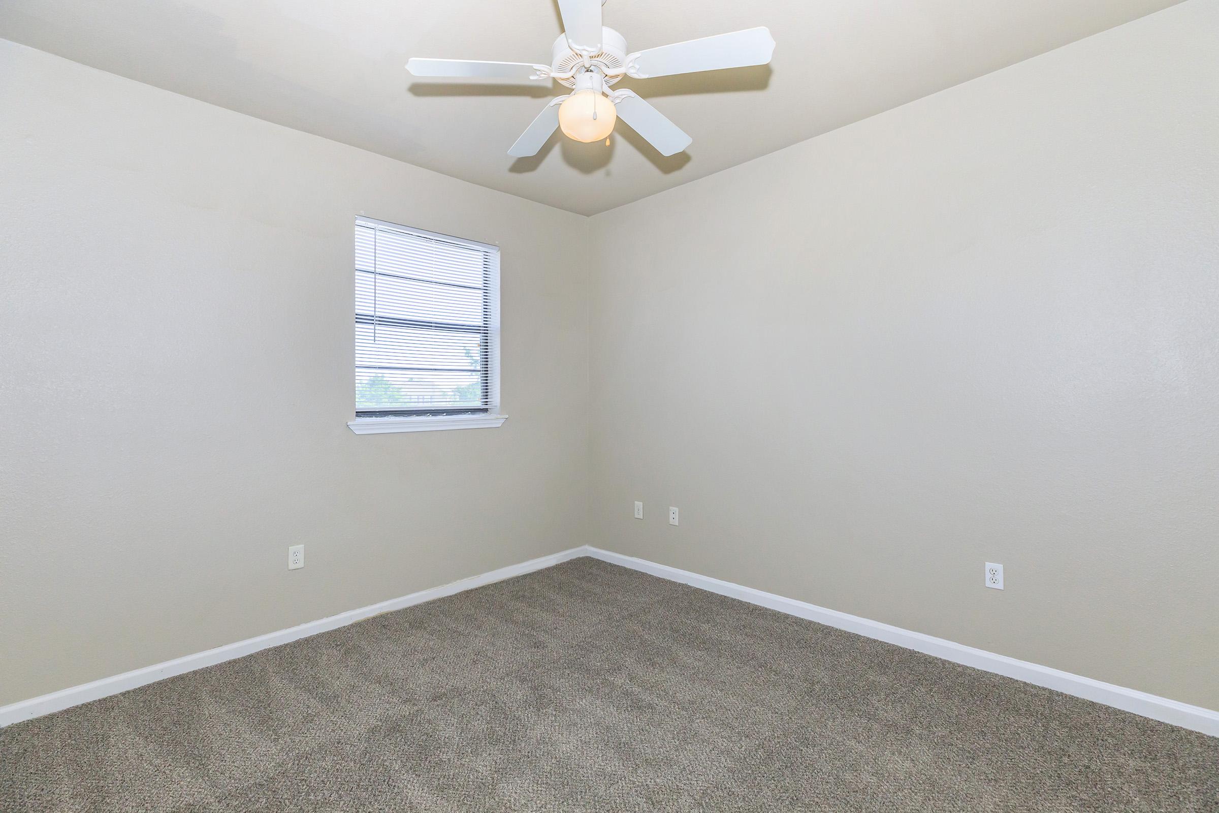 A spacious, empty room with light beige walls and gray carpet. The room features a ceiling fan with a light fixture and a single window with horizontal blinds, allowing natural light to enter. The overall atmosphere is clean and inviting, suitable for various uses.