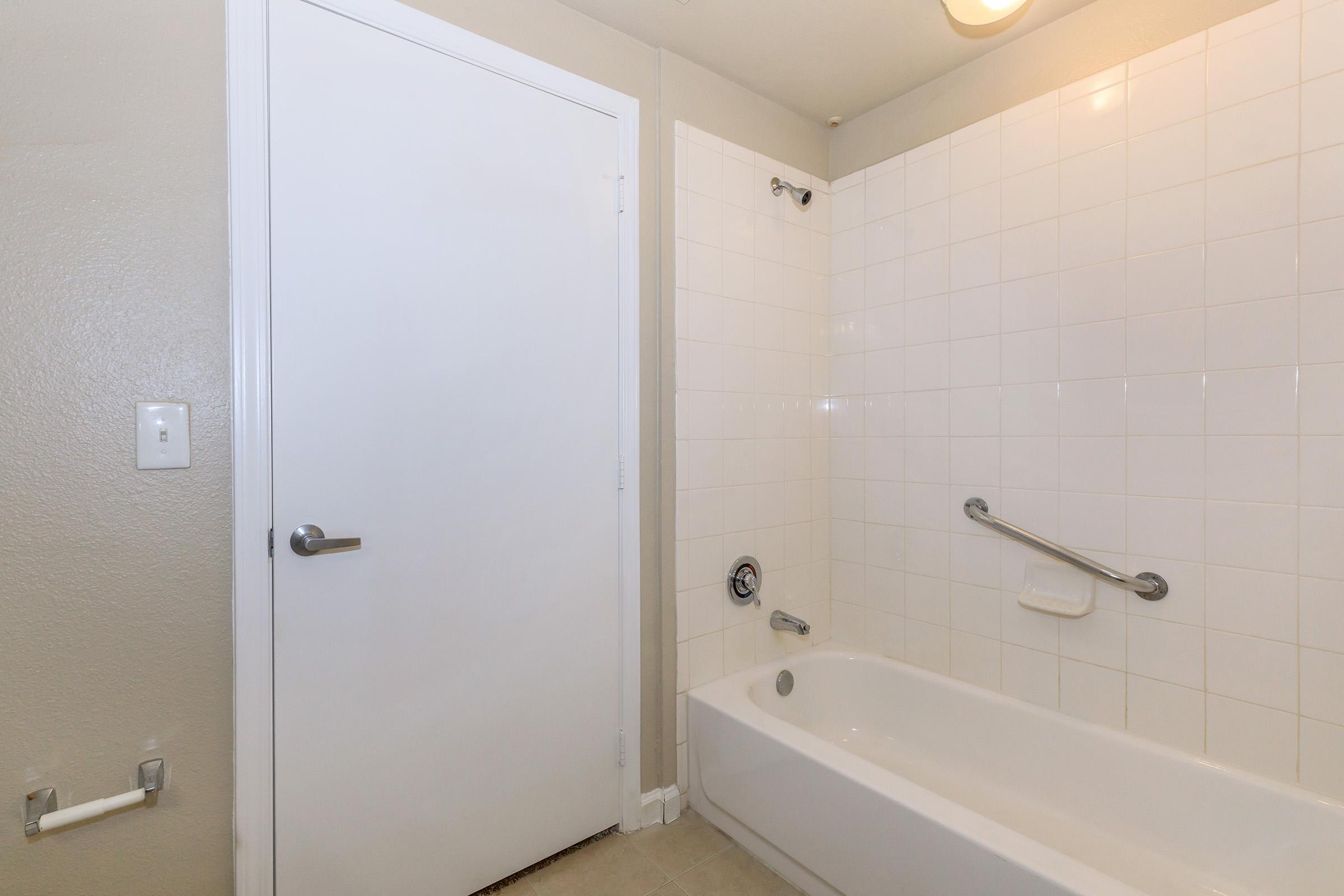 A clean, simple bathroom featuring a white tiled shower area with a bathtub, a white door leading to another room, and a grab bar on the wall for safety. The walls are painted in a light neutral color, providing a spacious and bright atmosphere.