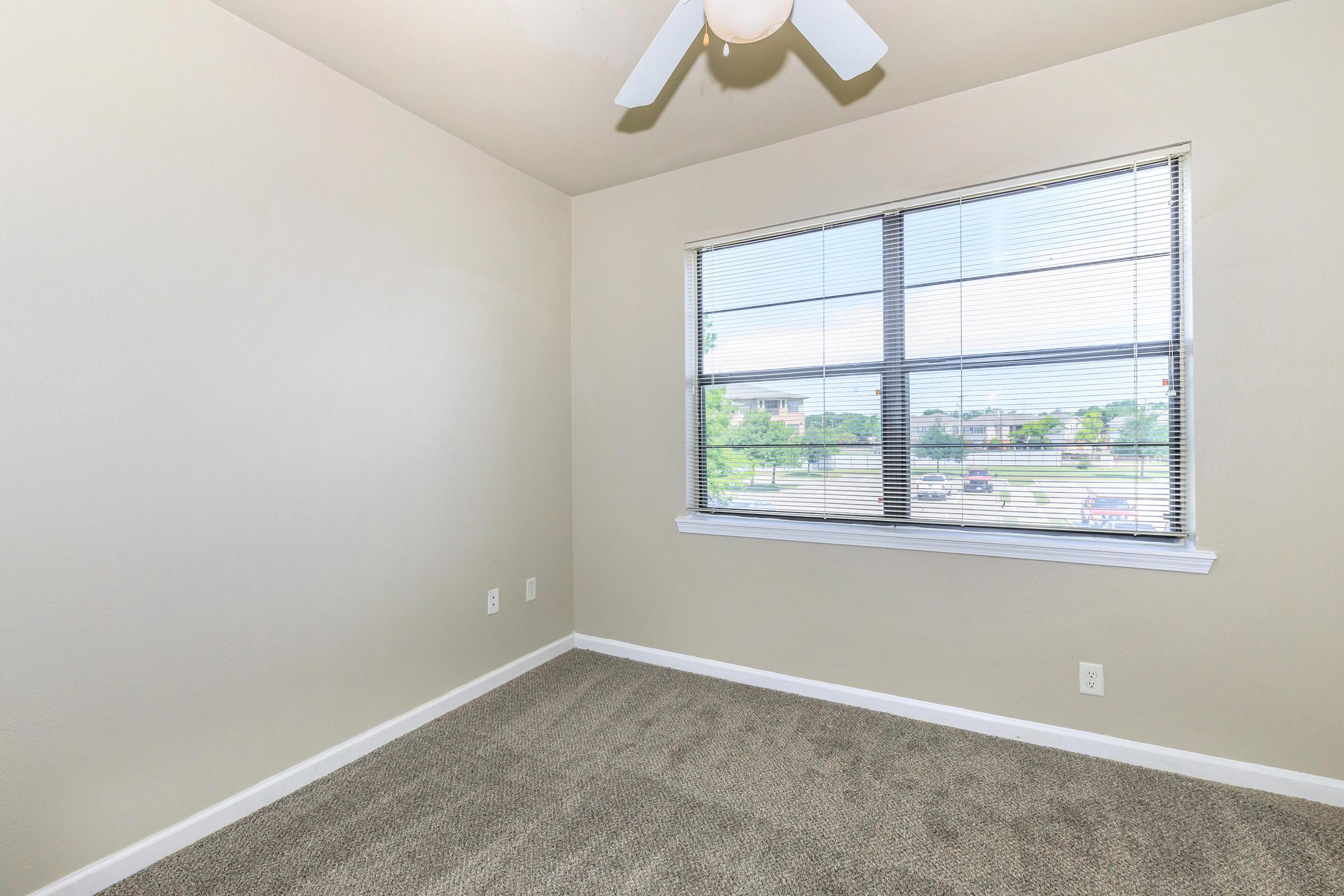 A vacant room with light beige walls and a ceiling fan. The floor is covered with carpet, and a large window with blinds allows natural light to enter, showcasing a view of greenery outside. The room is unfurnished, creating a spacious and inviting atmosphere.