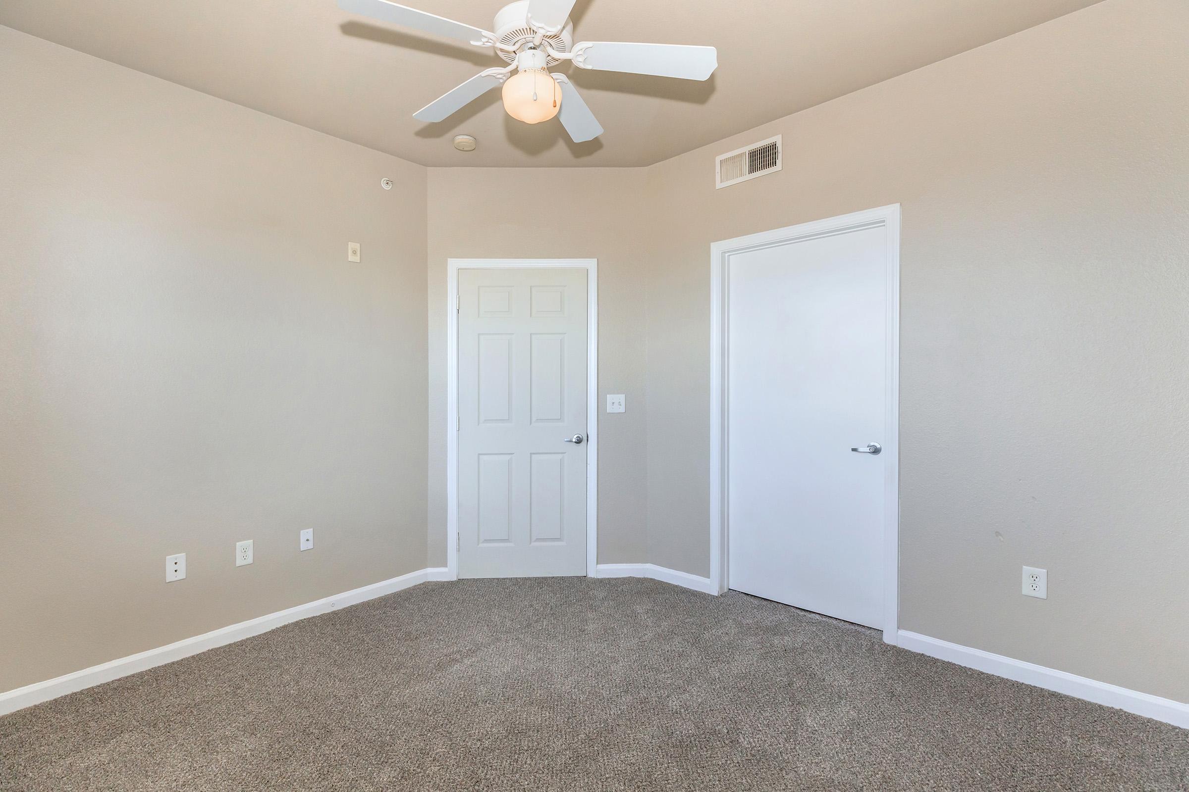 A neutral-toned room with light beige walls and plush carpet. It features a ceiling fan with five blades, two doors—one closed and one partially opened—and a small wall outlet. The room has no furniture, creating a spacious feel.