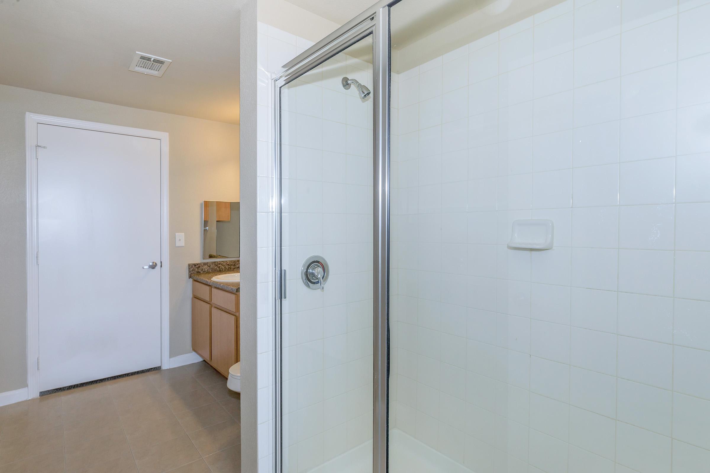 A modern bathroom featuring a glass shower enclosure and a doorway leading to another room. The space is well-lit, with beige walls and a tiled floor. A countertop with a sink is visible in the background, along with a mirror, creating a clean and functional design.