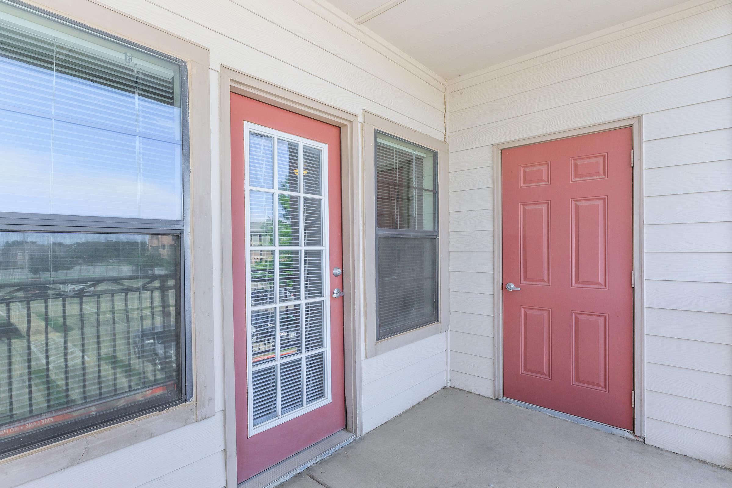 Two red doors on a light-colored exterior of a building. One door features a window with multiple panes, while the other is solid. There are large windows beside each door, allowing natural light to enter the area. The ground is concrete, and the overall setting appears to be a residential entrance or balcony.