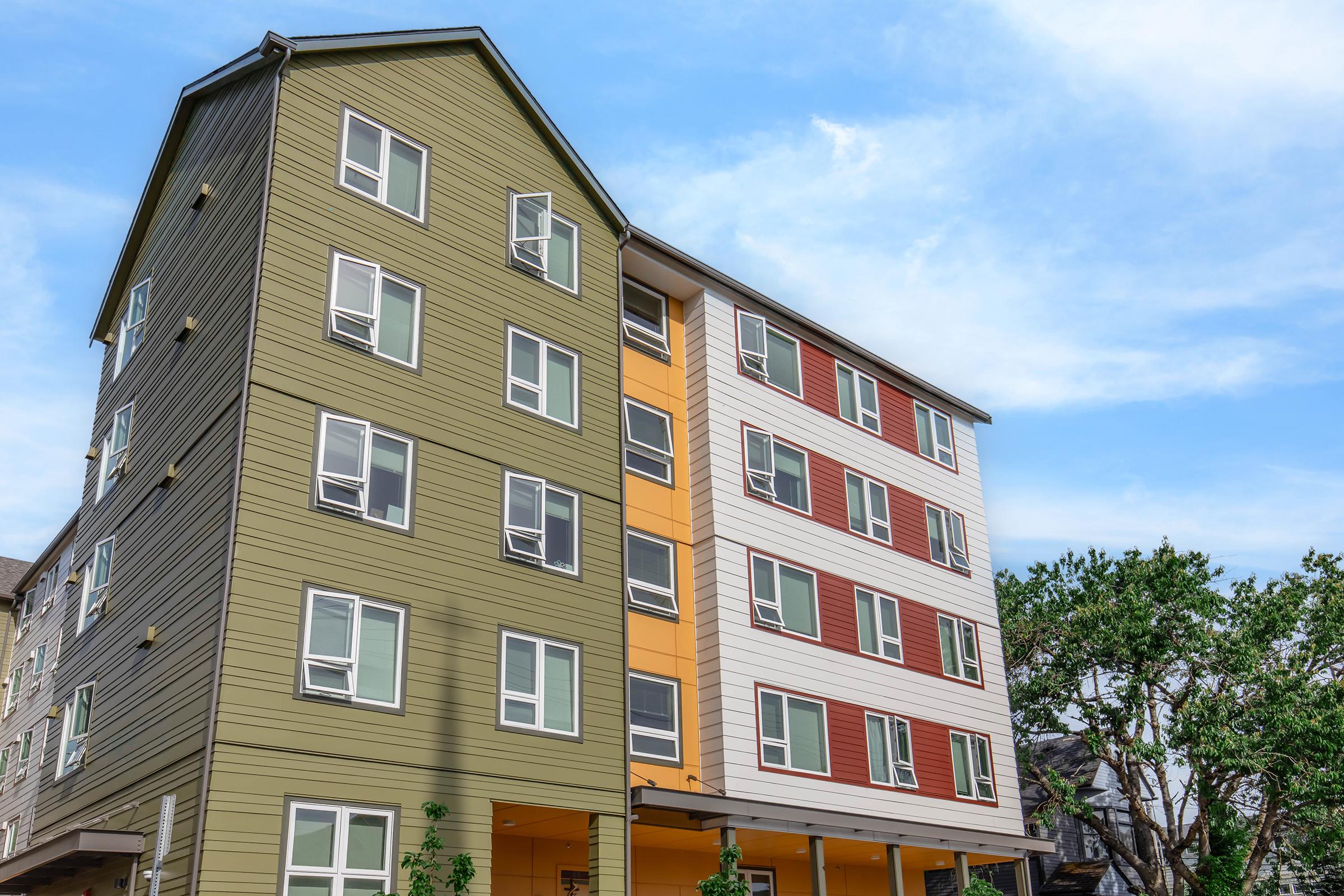A modern multi-story building featuring a mix of green, orange, red, and white wooden siding. The structure has large windows and is set against a bright blue sky with a few trees partially visible beside it.