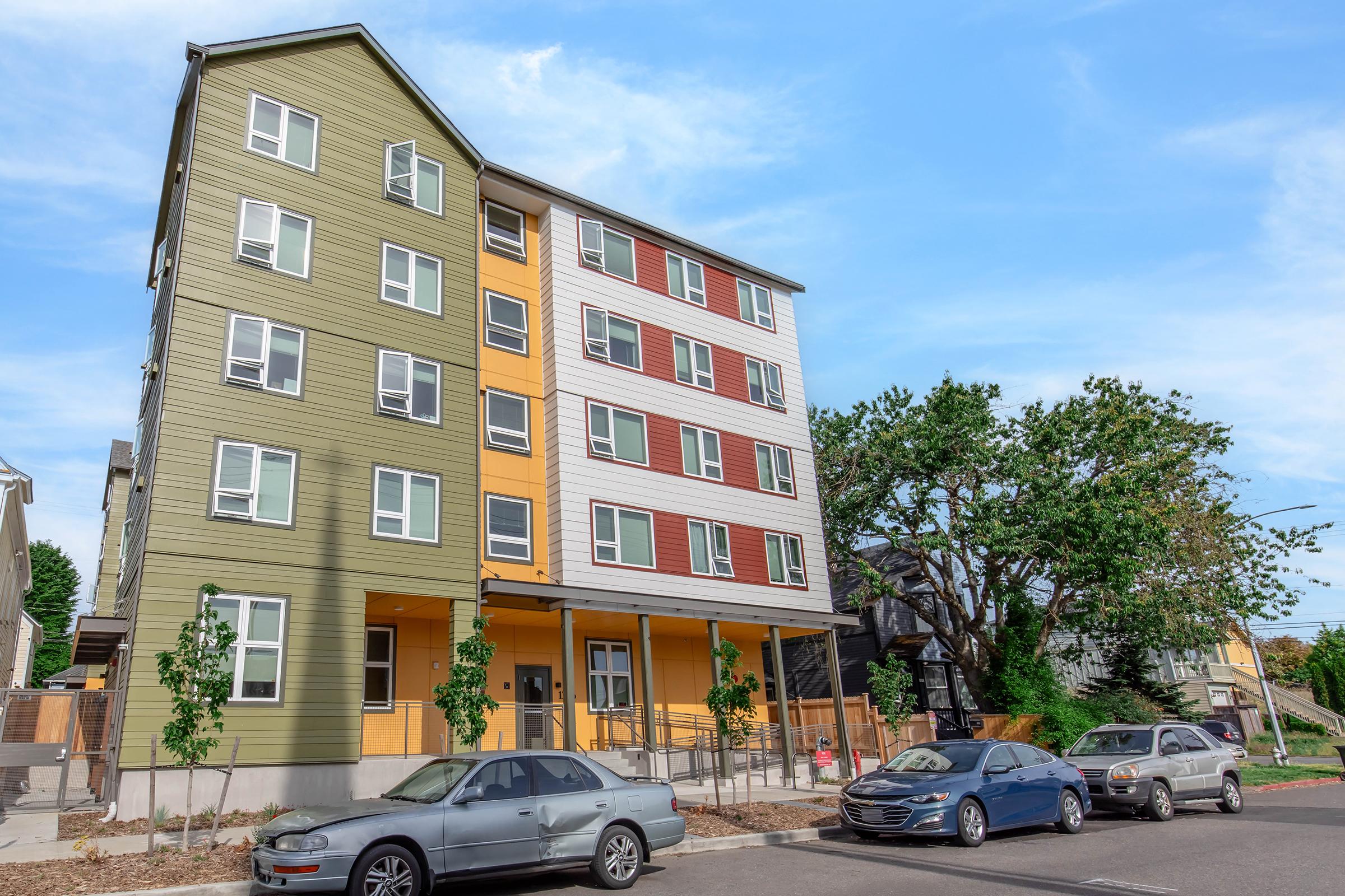 A multi-story modern apartment building featuring a mix of green, yellow, and red siding. Trees and landscaped areas are visible in the foreground, with parked cars along the street. The sky is clear and blue, creating a bright and inviting atmosphere.