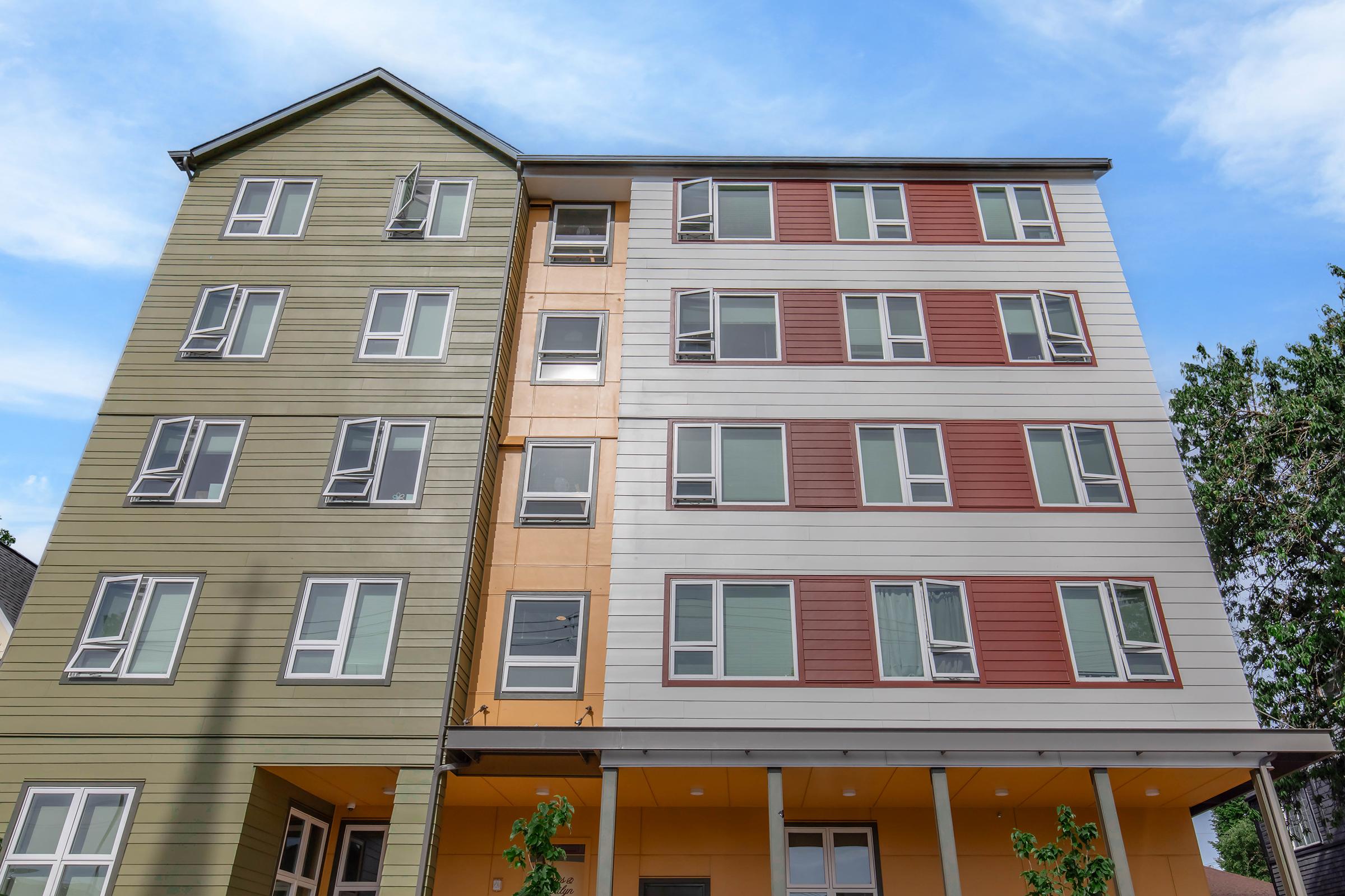 A modern multi-story building featuring a mix of green, orange, and red exterior panels. The façade has several windows, with some partially open. The structure is set against a clear blue sky, and small trees are visible at the base. The design appears contemporary and inviting.