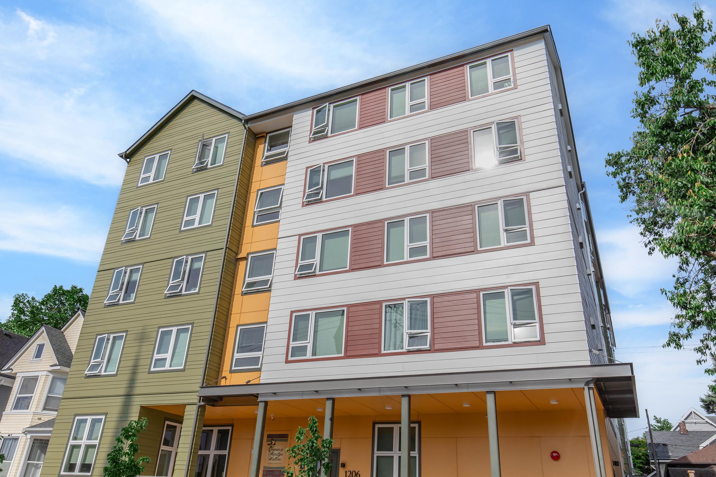 A modern multi-story building featuring a mix of exterior colors, including green, orange, and beige. The structure has large windows and a contemporary design, surrounded by trees and other residential buildings under a clear blue sky.