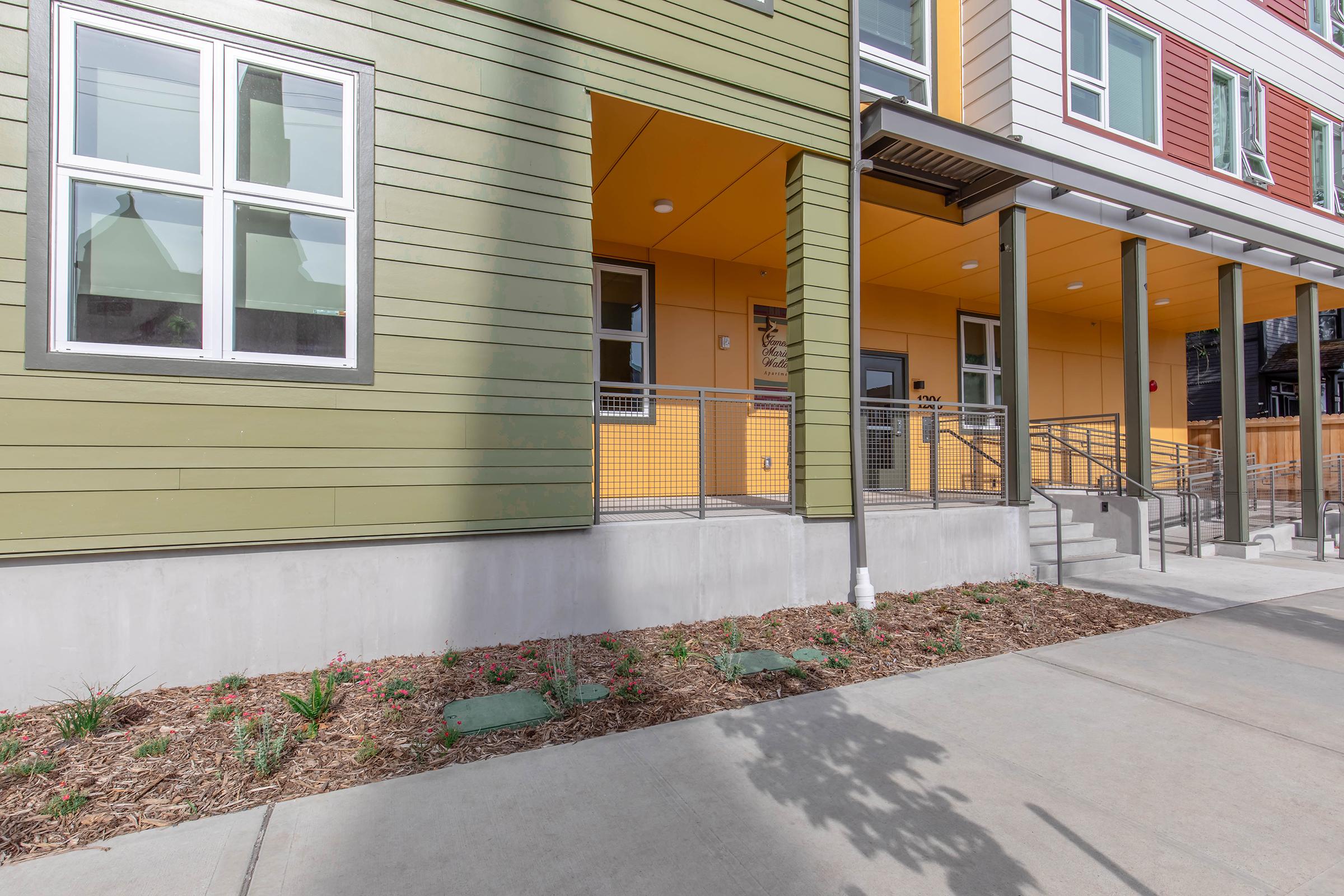 A modern building facade featuring green and yellow panels, large windows, and a front entrance with steps and a railing. The surrounding area includes a concrete walkway and landscaped garden beds with small plants, adding greenery to the urban setting. The sky is clear and bright.