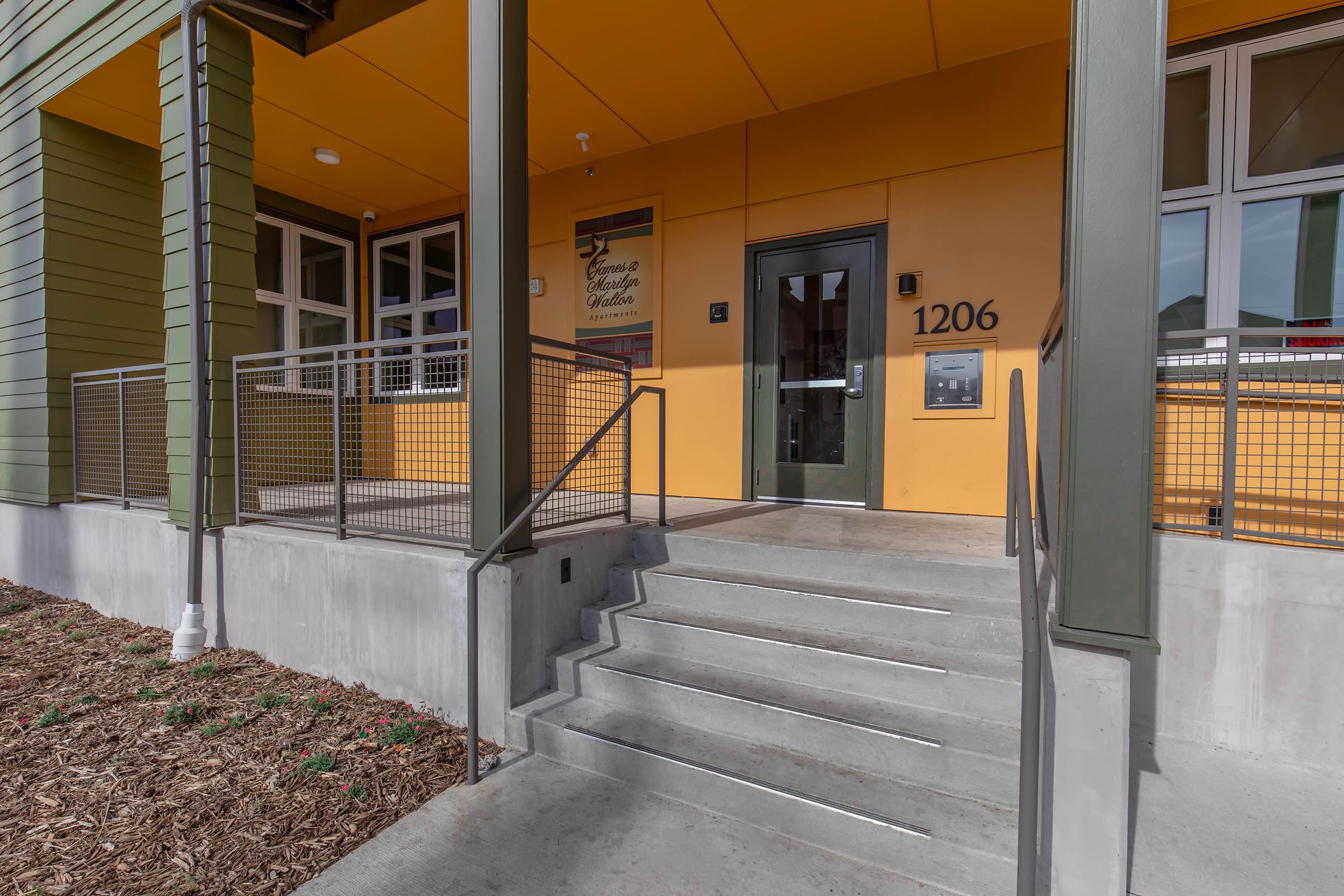 Front entrance of a building featuring a modern design with a yellow wall, a large door labeled "1206," a mailbox beside the door, and stairs leading up to the entryway. The area is landscaped with wood chips, and the entrance is framed by green walls and large windows.