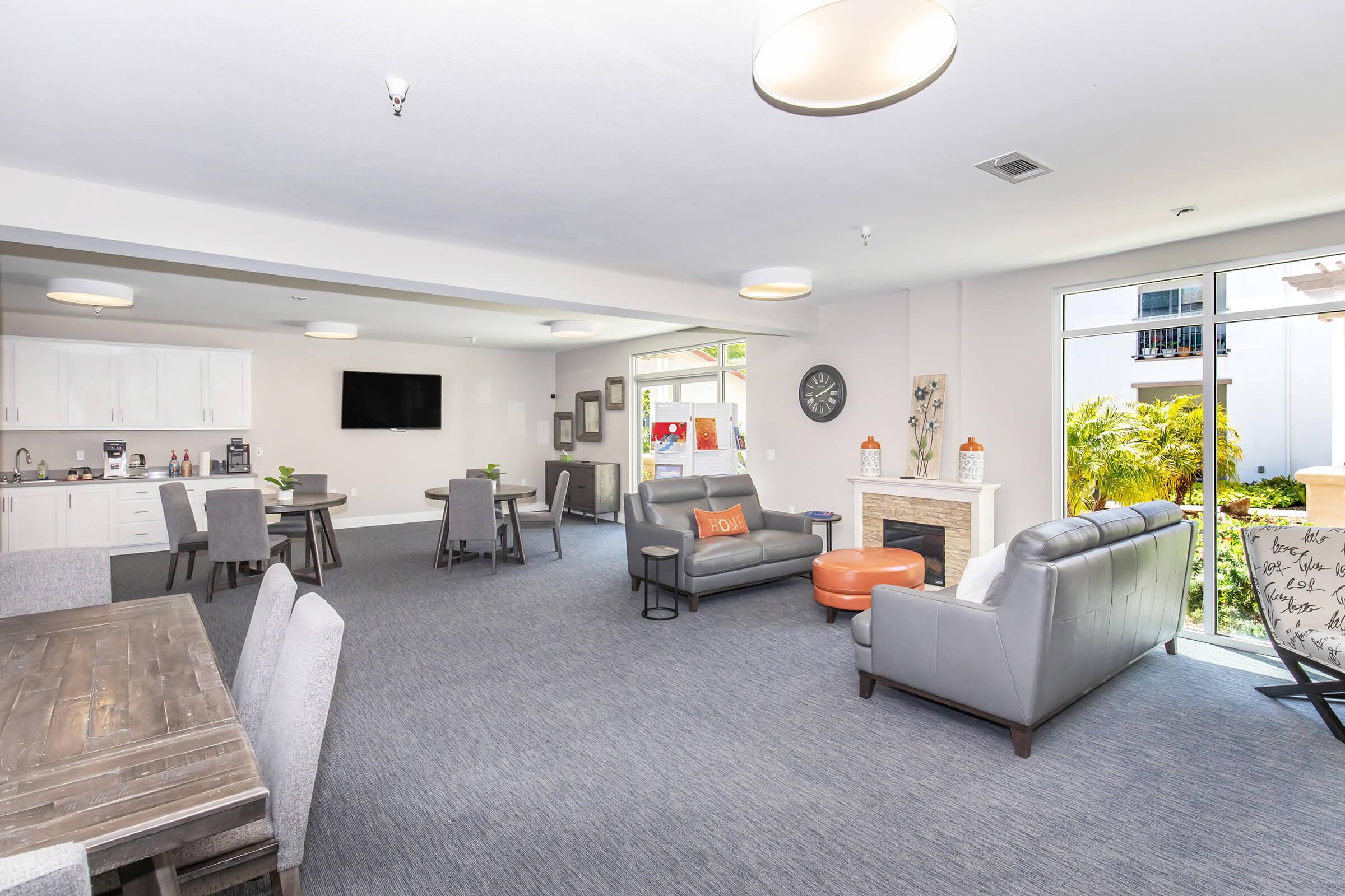 A modern communal living space featuring a seating area with gray sofas, a coffee table, and a large TV mounted on the wall. There are dining tables and chairs, a kitchenette with white cabinetry, and large windows that let in natural light. Decorative elements include plants and wall art.