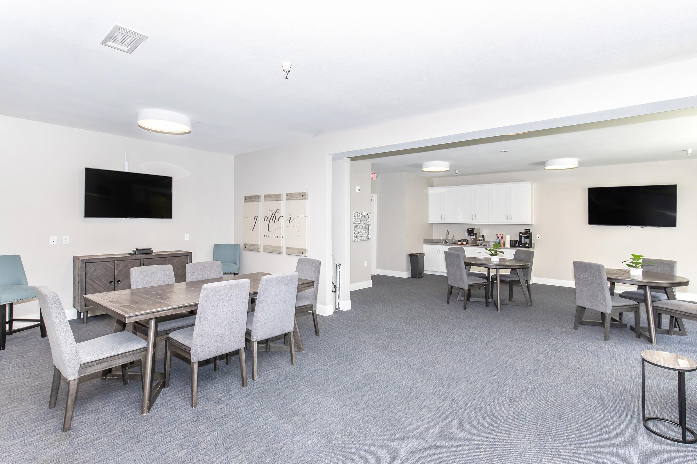 A spacious common area featuring multiple tables and chairs, with light gray upholstered furniture. Two flat-screen televisions are mounted on the walls. A kitchenette with white cabinets and modern appliances is visible in the background. The floor is covered with blue carpet, creating a cozy atmosphere.