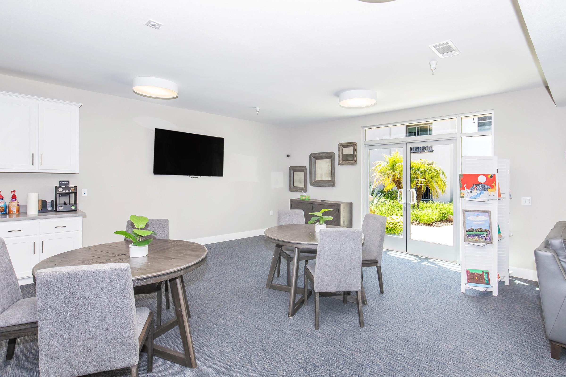 A modern common area featuring two round tables with four gray chairs each, a television on the wall, a small kitchenette area with coffee and snacks, and large windows that offer a view of greenery outside. The space is brightly lit and designed for comfort and social interaction.