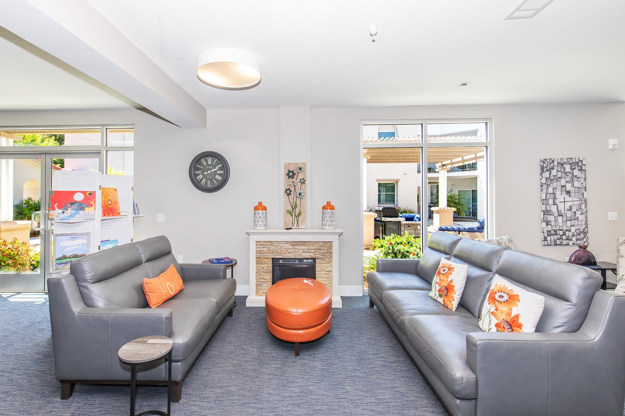 A modern living room featuring two gray sofas with orange pillows, a round orange ottoman, and a stone fireplace. Large windows allow natural light to fill the space, revealing an outdoor area with greenery. A decorative wall clock and artwork add character to the room.