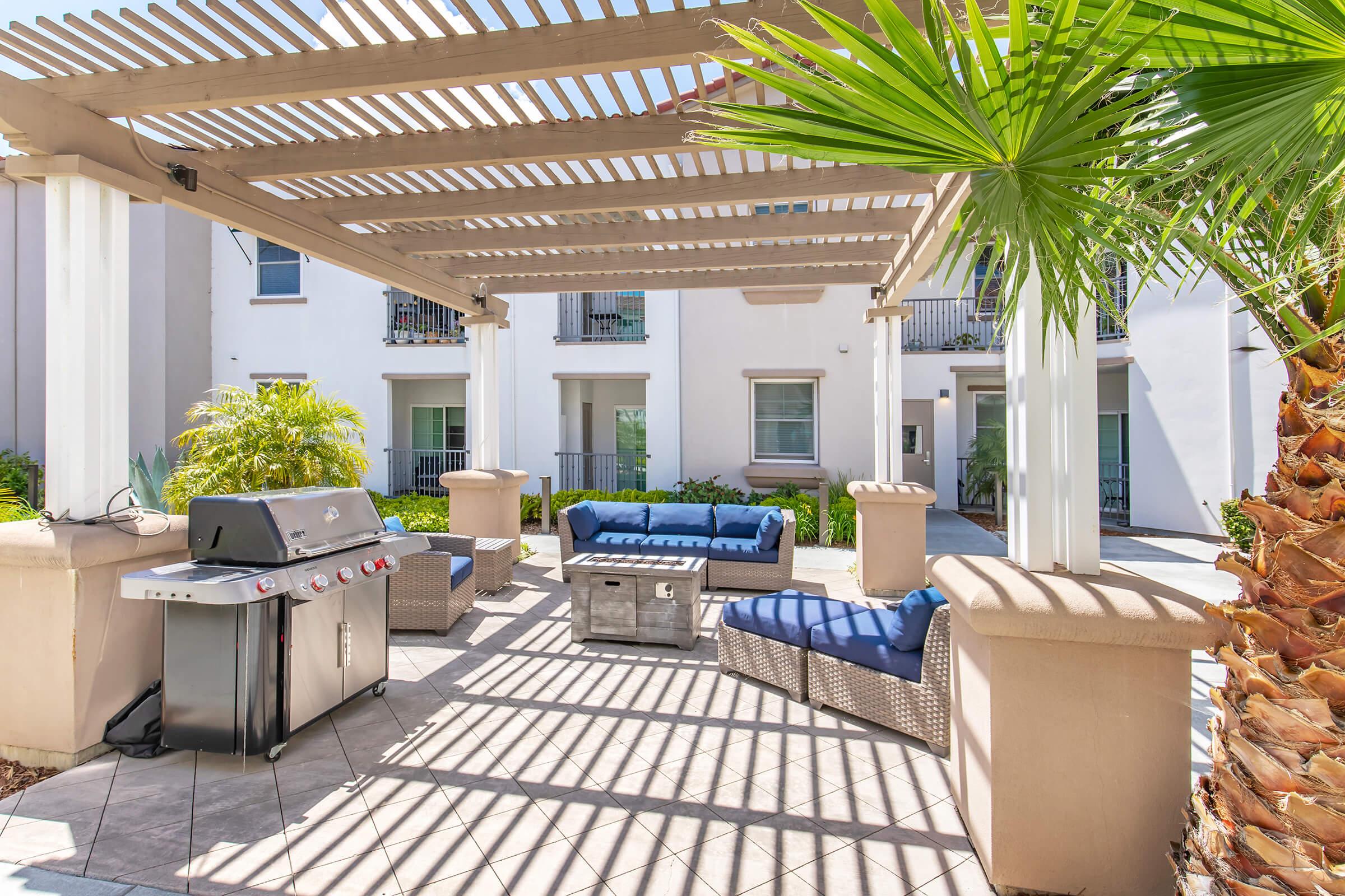 Outdoor patio area featuring a shaded seating arrangement with blue cushions, a coffee table, and a gas grill. Surrounded by lush greenery and palm trees, the space is designed for relaxation and entertainment. Sunlight creates a pattern of shadows on the tiled floor.