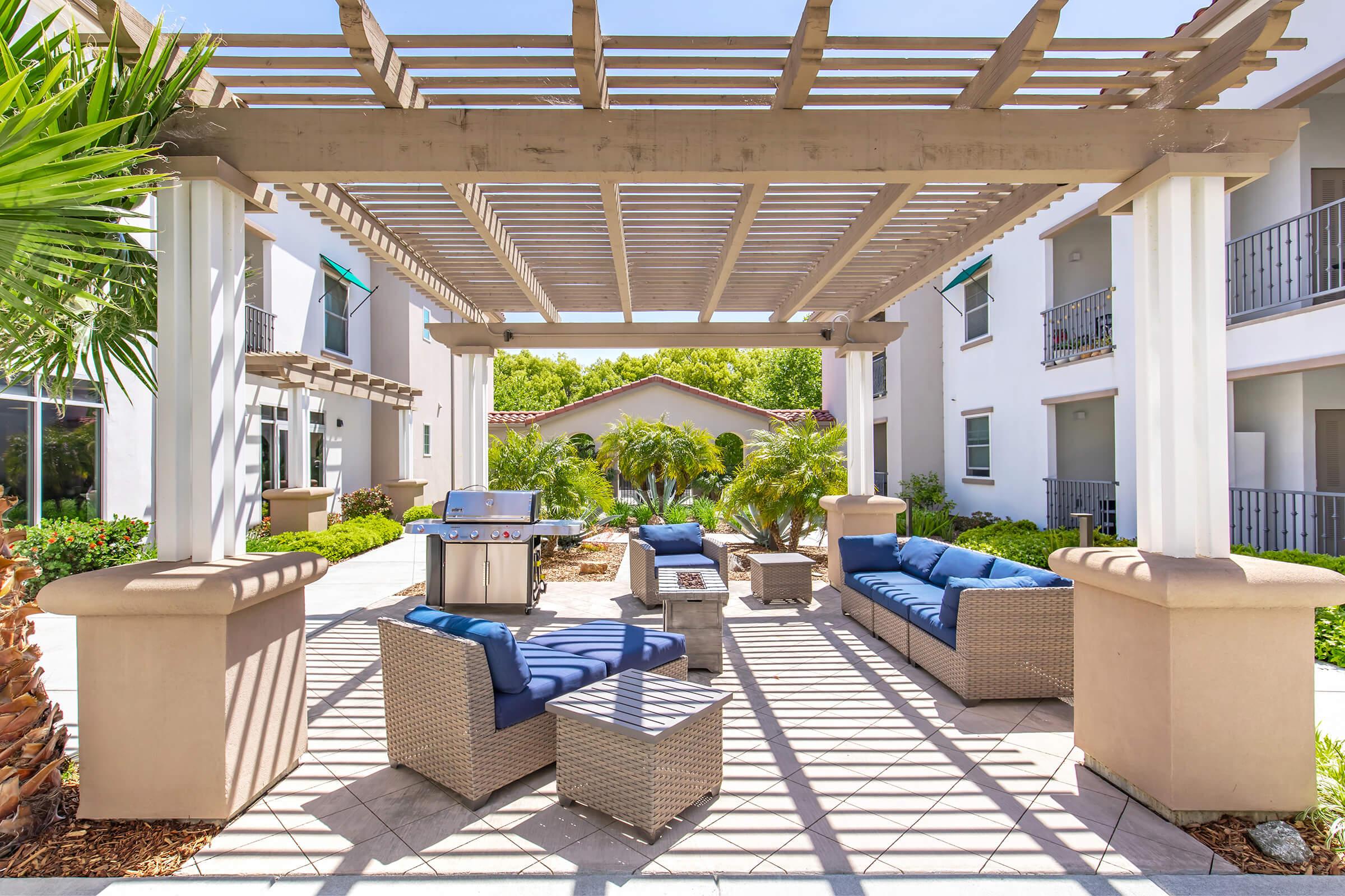 Outdoor lounge area featuring comfortable blue seating beneath a pergola, with a fire pit and a grill nearby. Surrounding greenery adds a touch of nature to the space, which is part of a well-maintained building. Bright sunlight casts shadows on the patio.