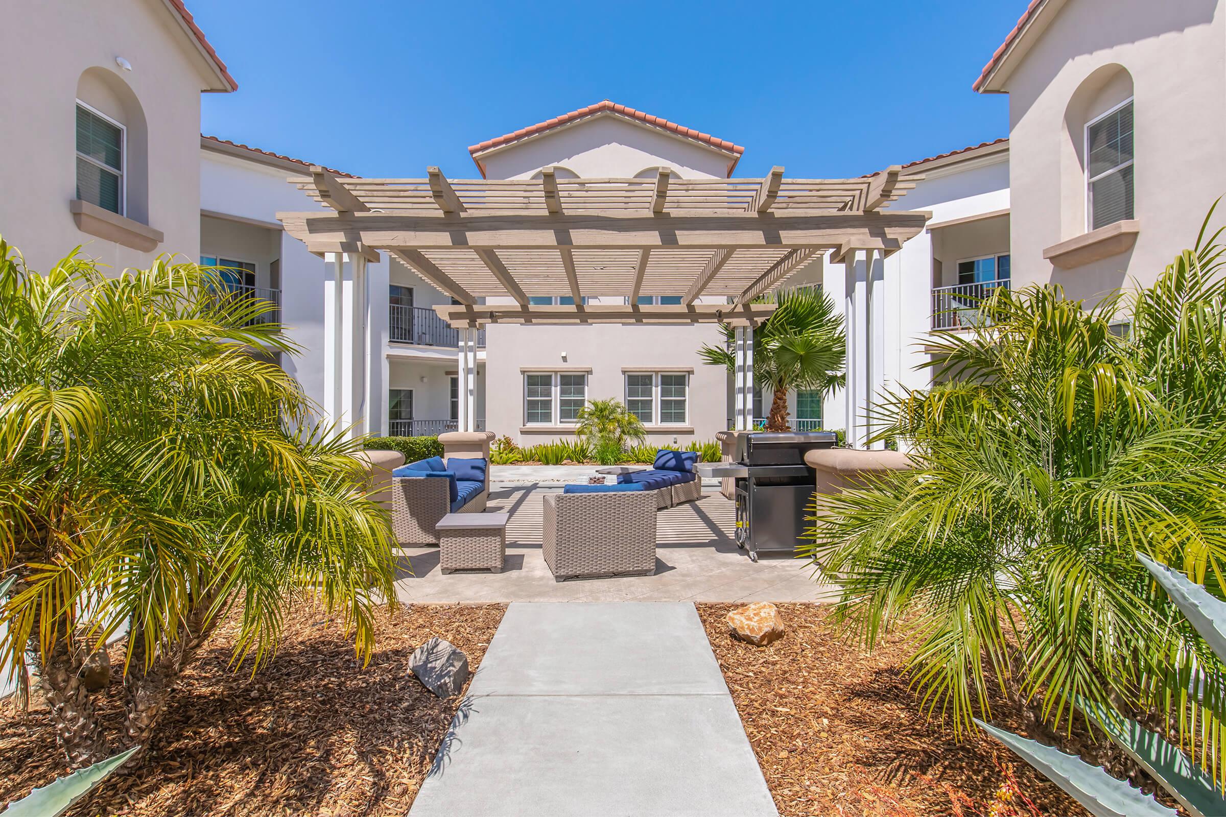A well-decorated outdoor common area featuring a pergola, comfortable seating, and a barbecue grill. Lush palm trees and other greenery surround the space, providing a relaxing atmosphere. The blue sky and sunlight enhance the cheerful ambiance of this residential community.