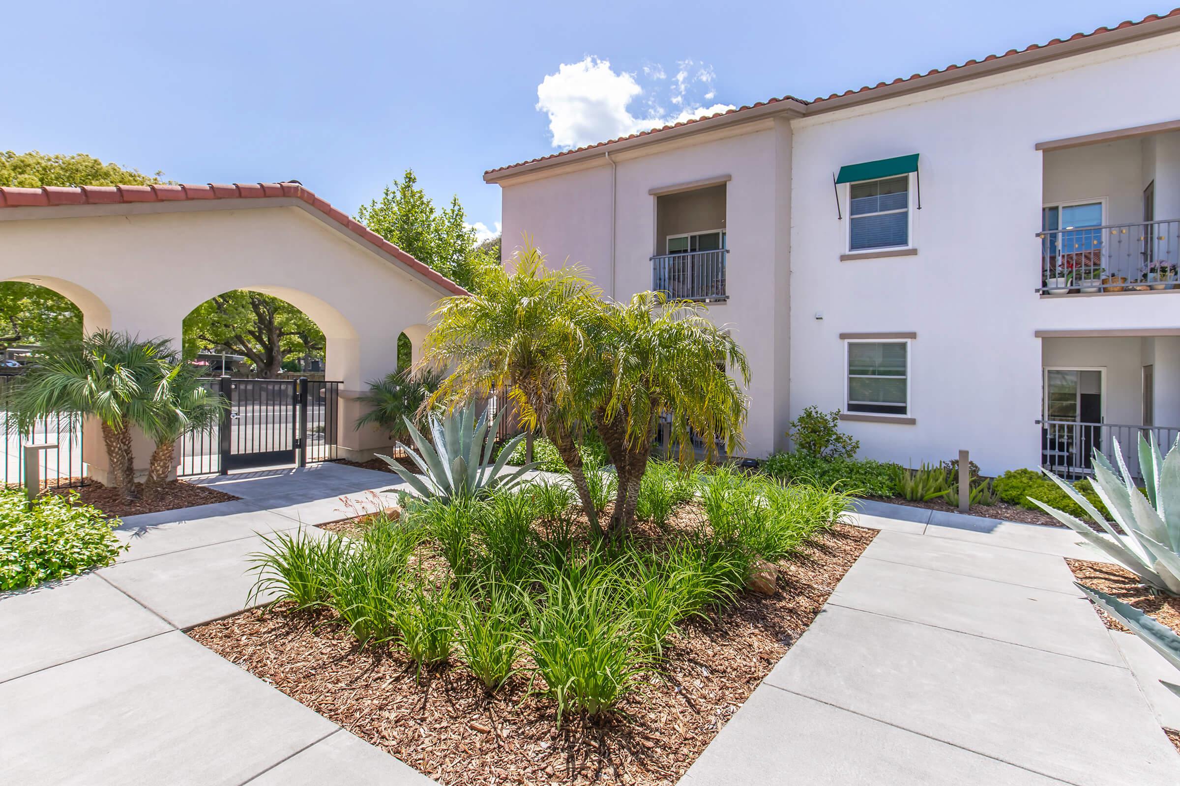 Lush courtyard with green plants and palm trees, featuring concrete pathways. The scene includes a gated entrance with an arched structure on the left and a two-story building with balconies on the right, under a blue sky with some clouds.