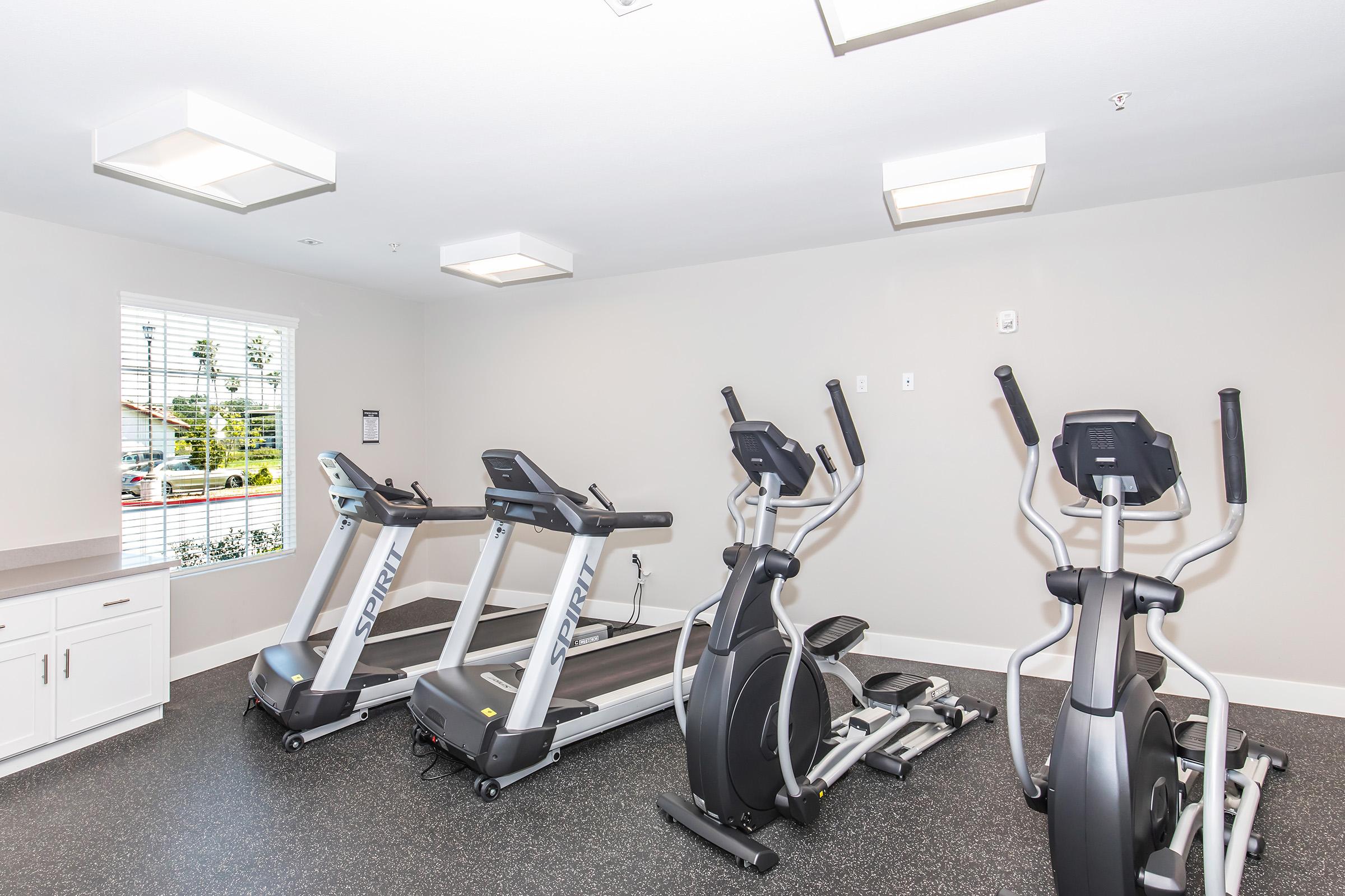 A modern gym interior featuring multiple cardio machines, including treadmills and ellipticals, on a rubberized floor. Natural light streams through a window, complemented by bright overhead lighting. The walls are painted in a neutral tone, creating a clean and inviting workout space.