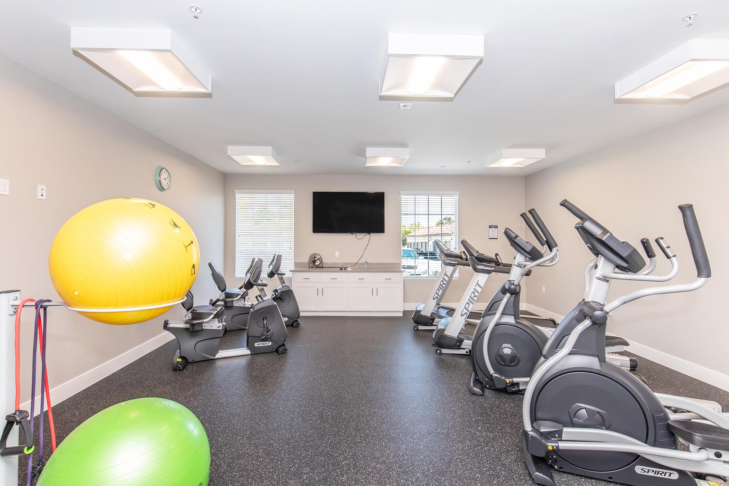 A modern gym interior featuring exercise equipment including stationary bikes and fitness balls. The room is well-lit with overhead lighting, and there is a wall-mounted TV. Large windows provide natural light, and the flooring is a black rubber material suitable for workouts.