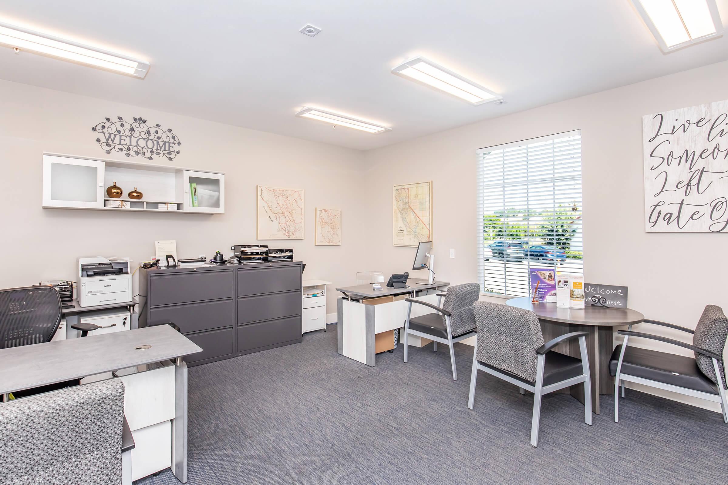 A modern office space featuring gray carpeting, two desks with chairs, a printer, and storage cabinets. The walls are adorned with artwork and a "Welcome" sign. Natural light streams through large windows, providing a bright and inviting atmosphere.
