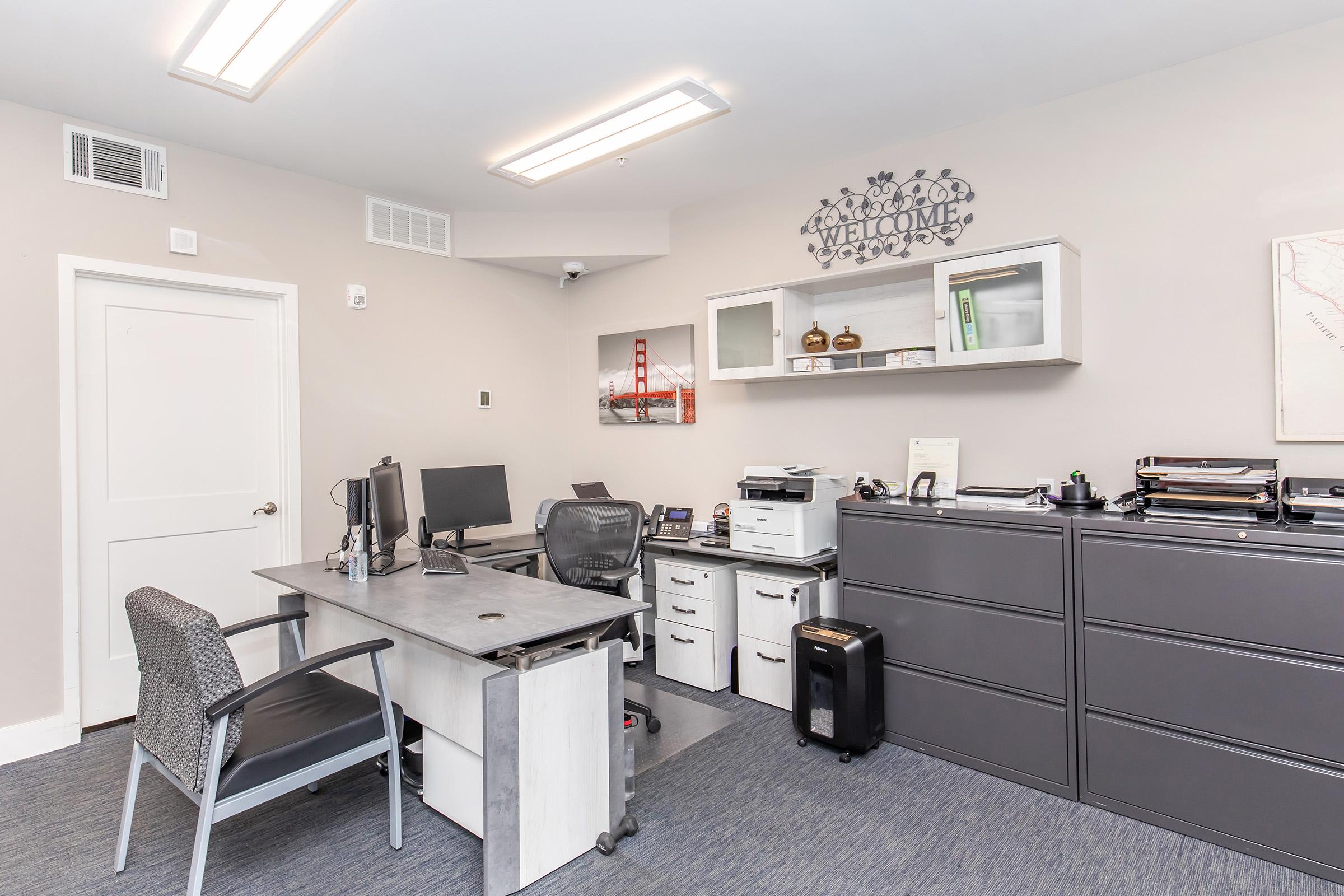 A modern office space featuring a desk with a computer and chair, a printer, filing cabinets, and decorative wall art. The room is well-lit with overhead lights and has a welcoming atmosphere, ideal for professional work.
