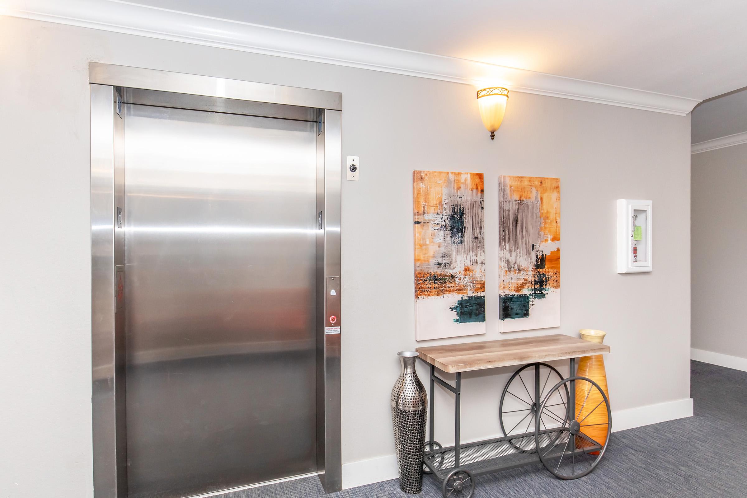 A modern elevator in a hallway with a neutral color scheme. Next to it is a wooden table with a decorative wheel, a tall vase, and abstract artwork on the wall featuring orange and gray tones. Soft lighting enhances the inviting atmosphere of the corridor.