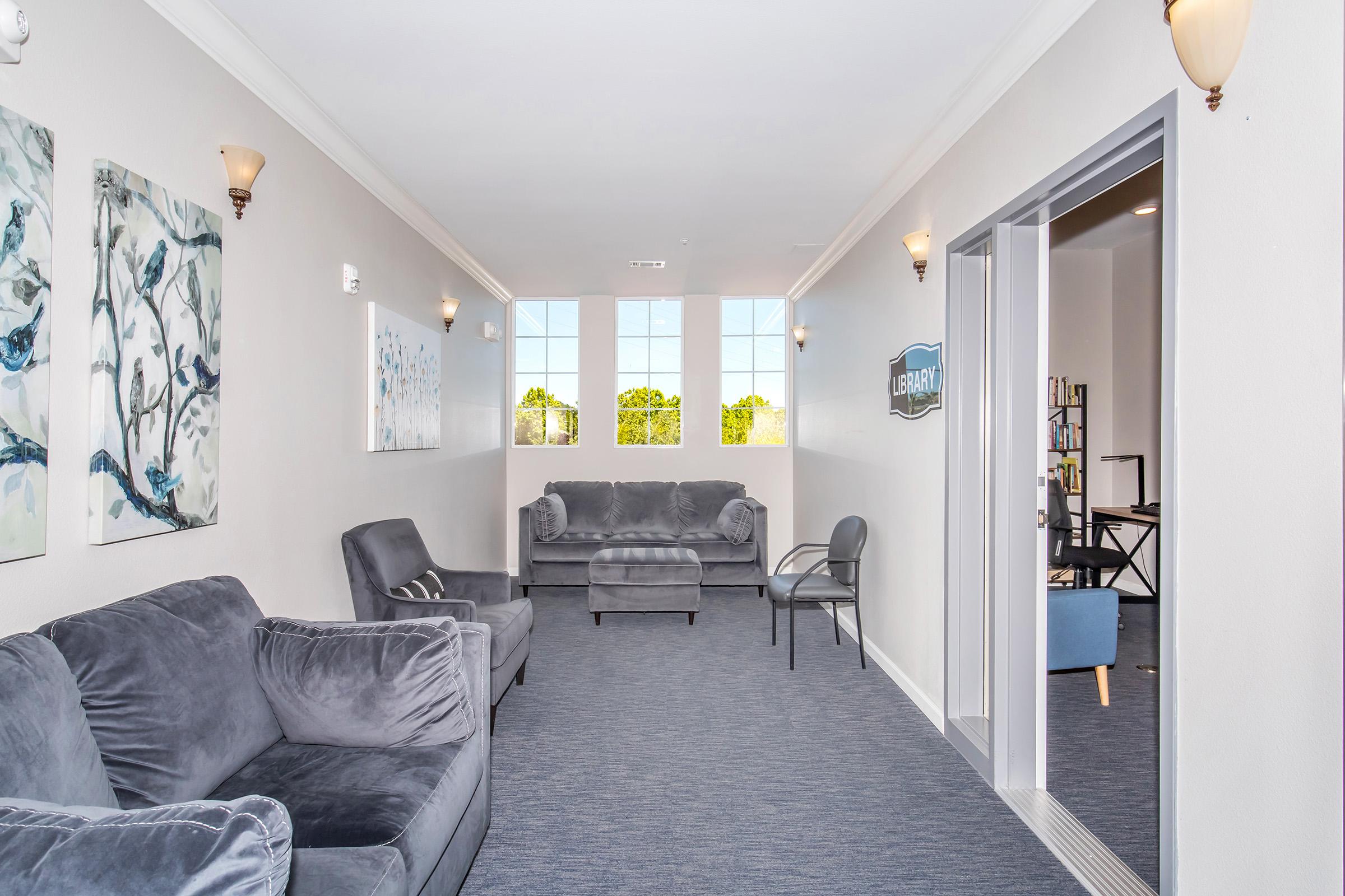 A cozy library hallway featuring two comfortable gray sofas, an armchair, and natural light from large windows. The walls are adorned with decorative artwork, and there is a sign indicating the entrance to the library. The flooring is carpeted, creating a warm and inviting atmosphere.