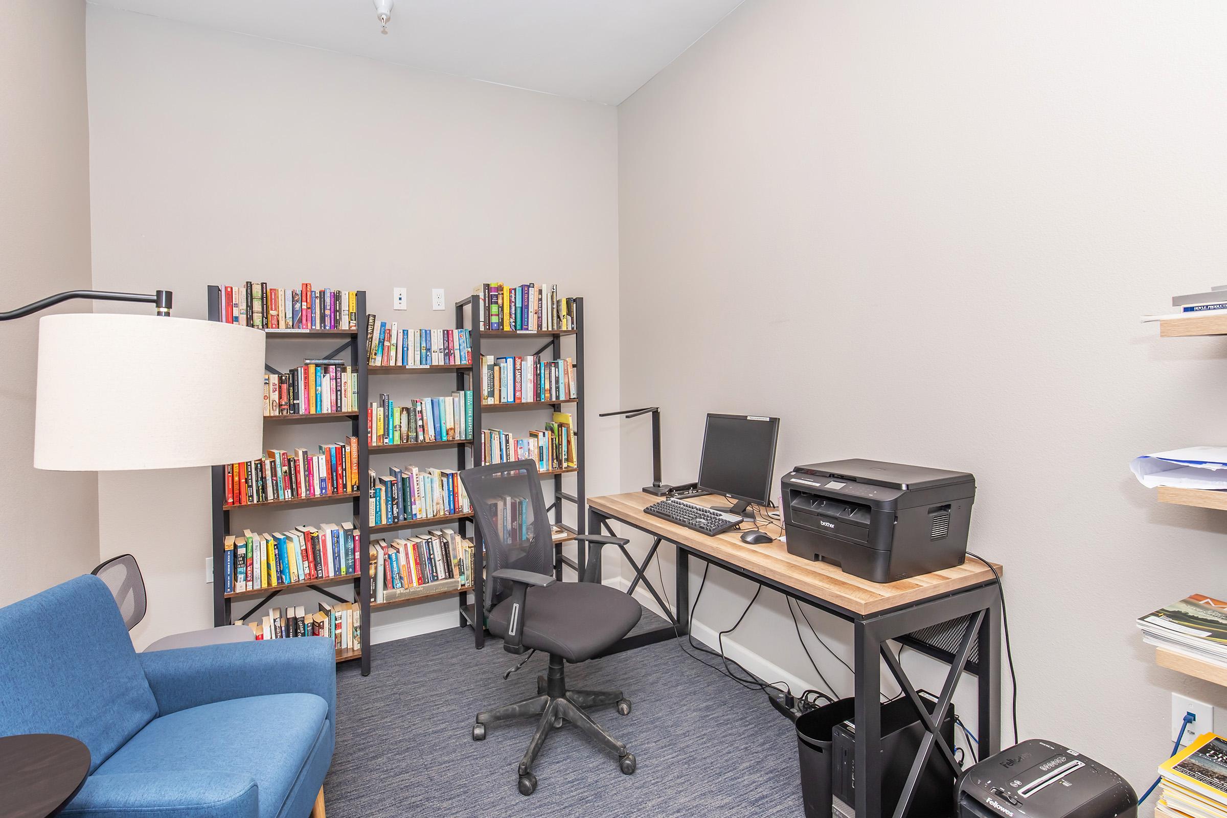 A small office space featuring a blue armchair, a black office chair, a wooden desk with a computer and printer, and two bookshelves filled with various books. The walls are painted in a neutral tone, creating a calm and organized environment suitable for reading or working.