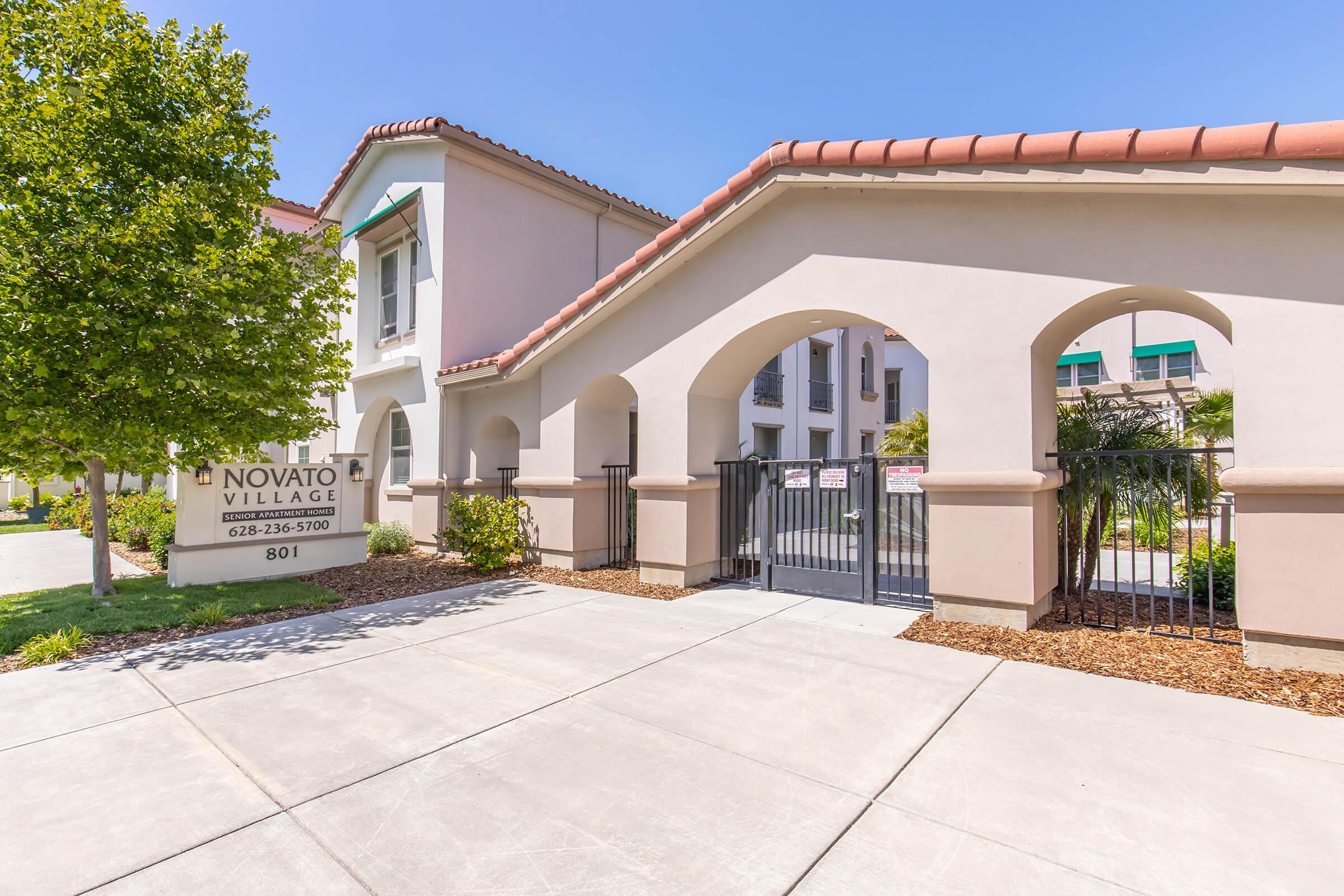 Entrance to Novato Village, featuring an archway with a gated entrance. There are landscaped areas with trees and a sign displaying the name "Novato Village" along with a contact number. The building has a light-colored exterior and a tiled roof, set against a clear blue sky.